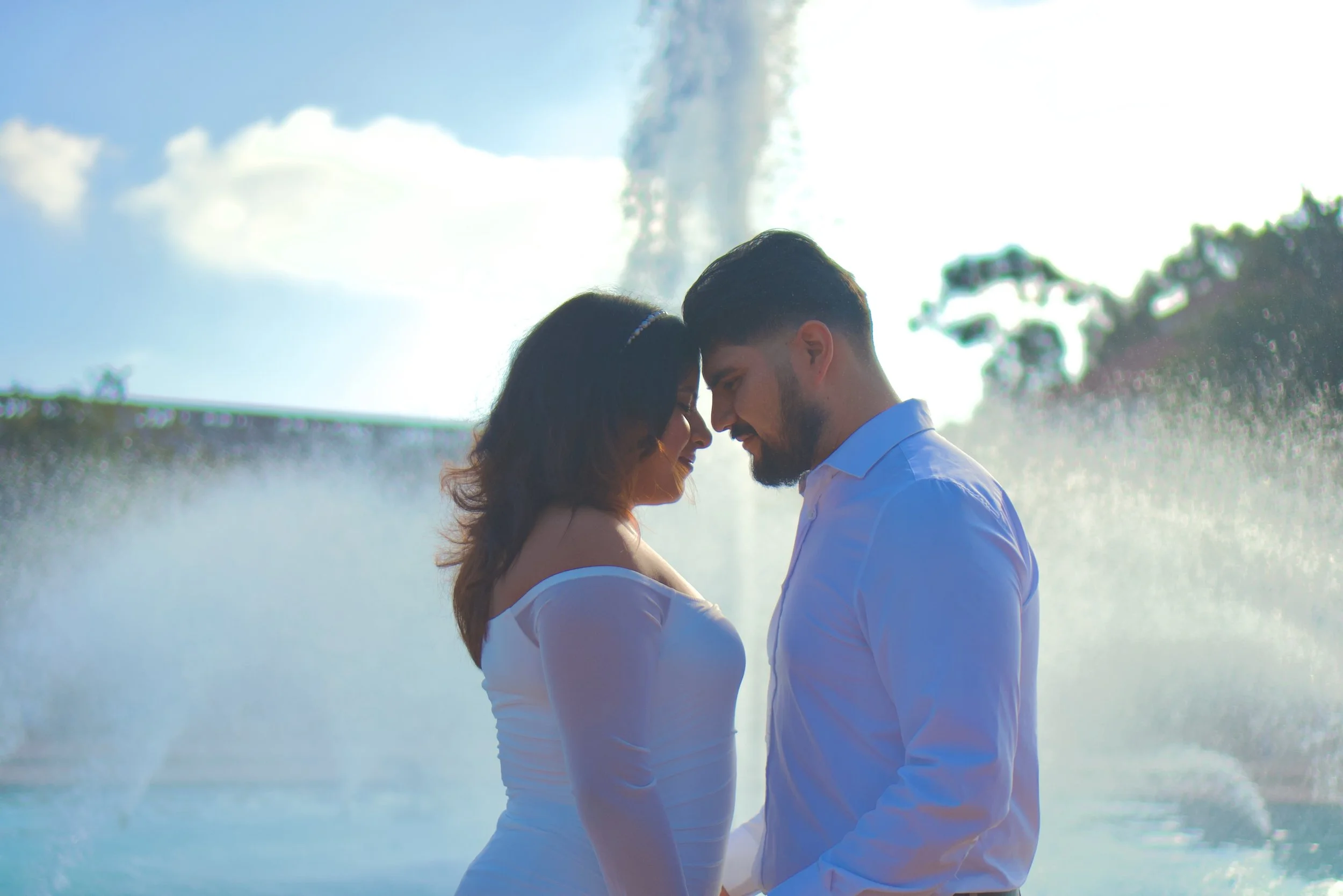 A couple with their foreheads touching, standing close in front of a large fountain with water spraying in the background, on a sunny day.
