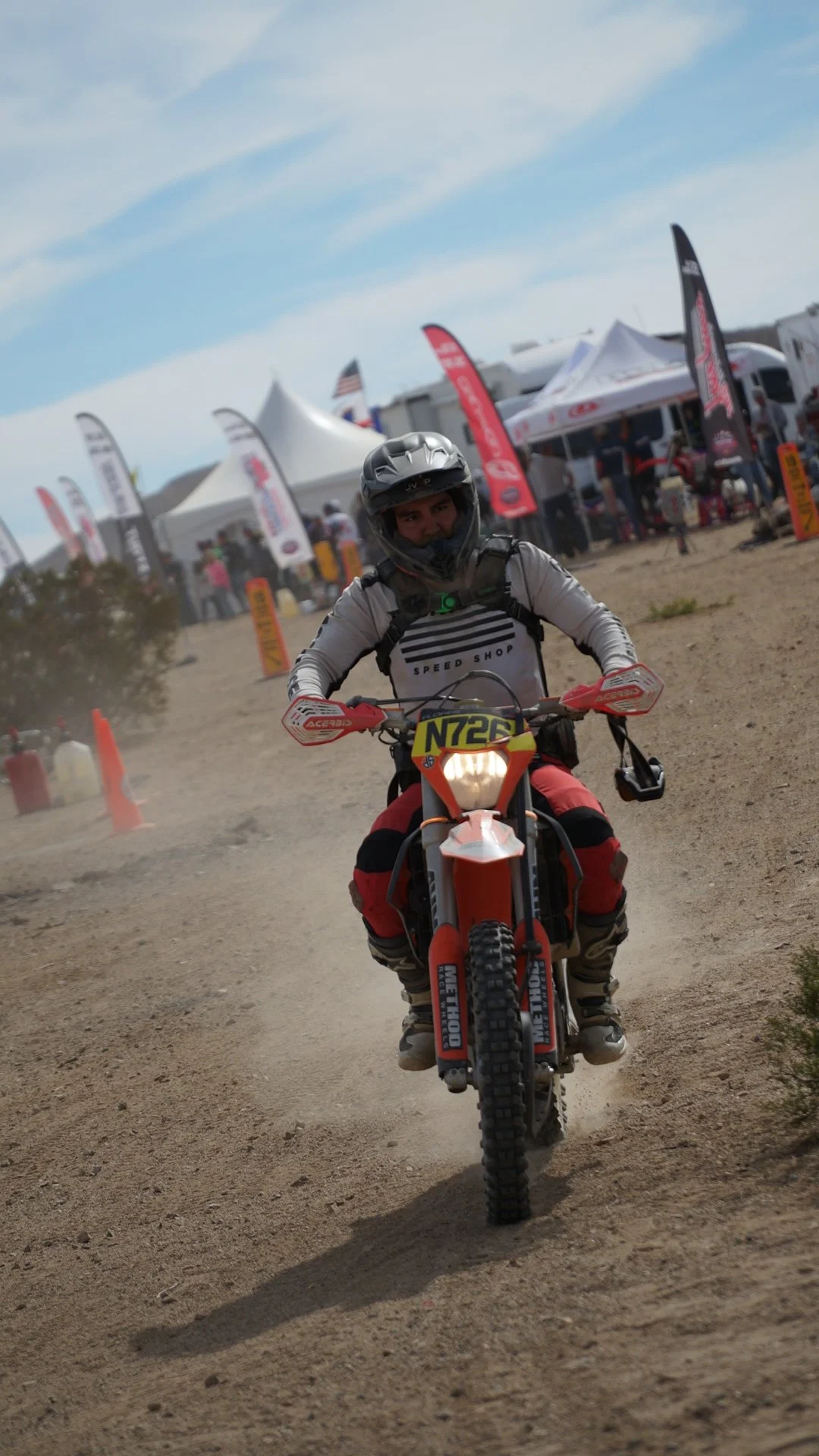 A person riding a dirt bike on a dusty trail at an outdoor event with tents and flags in the background.