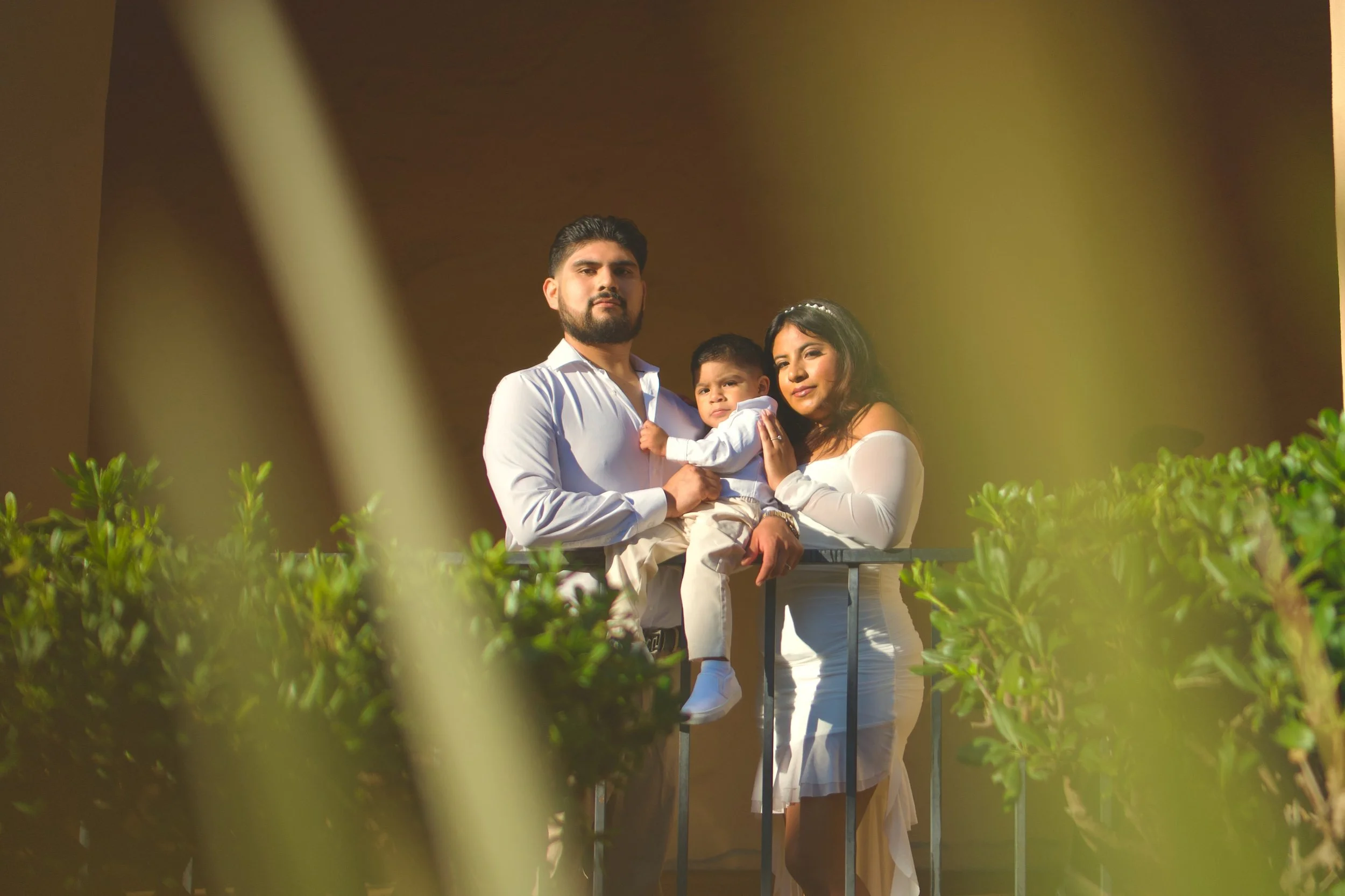 A family of three standing on a balcony, with the father holding a young boy and the mother standing next to them, surrounded by greenery.