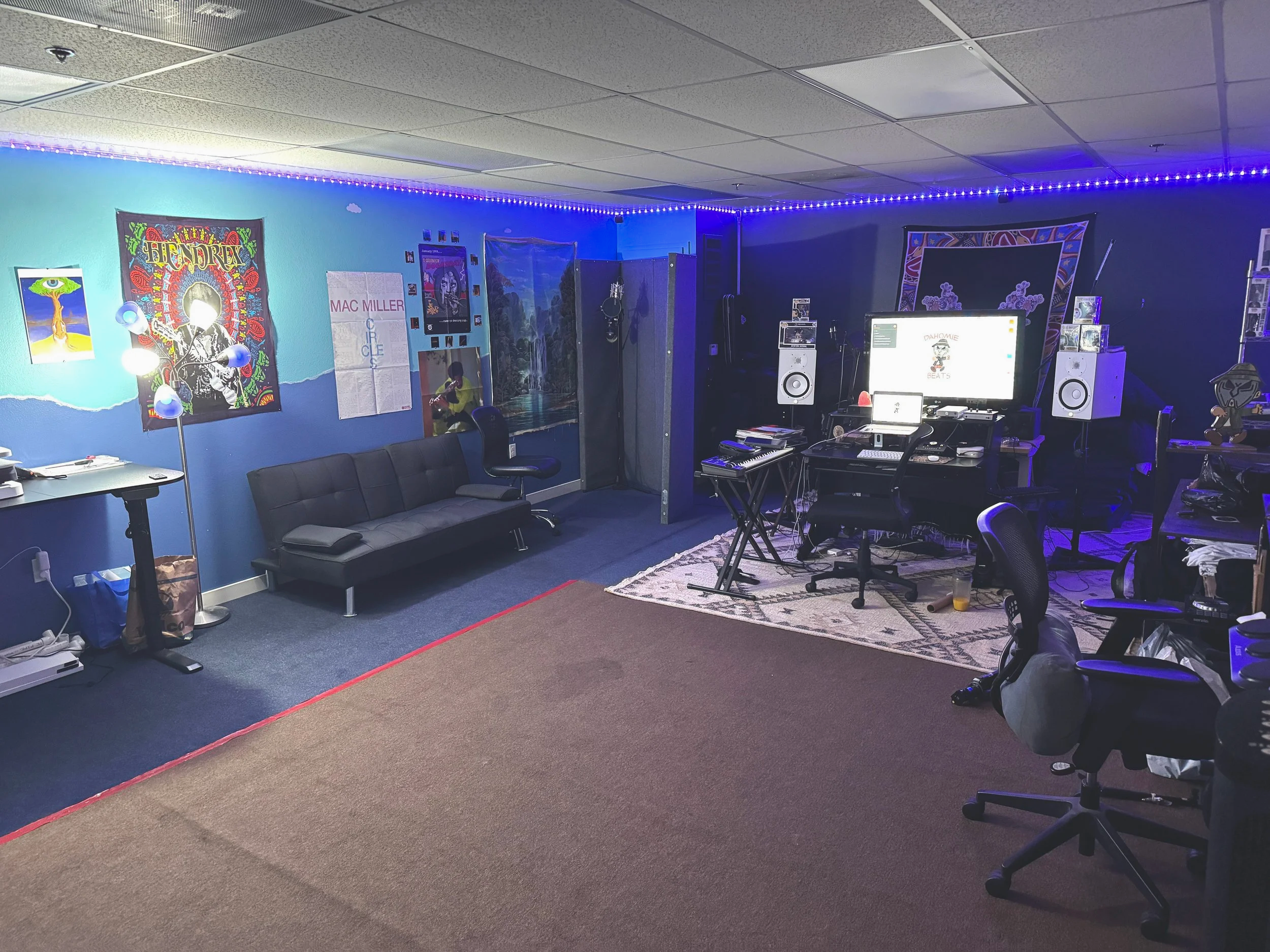 A room decorated with musical equipment, posters, and artistic decor featuring blue lighting along the ceiling. There are several chairs, a desk with a computer, and a black couch against a wall with paintings and posters. The space appears to be a creative or music studio.