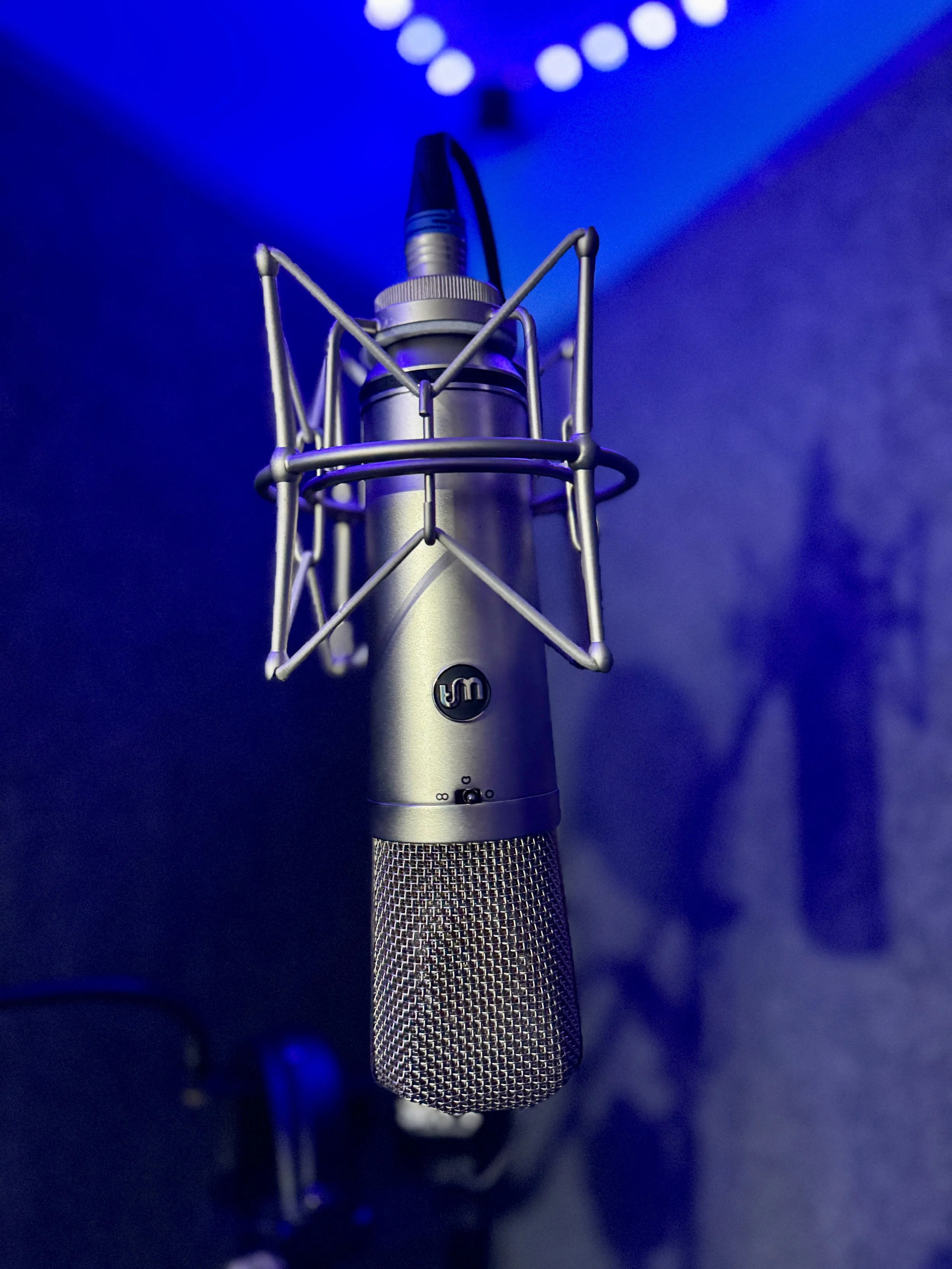 Close-up of a professional condenser microphone with a shock mount suspended in a recording studio with blue lighting.