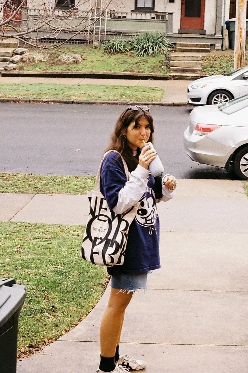 Young woman with glasses on her head, wearing a blue oversized T-shirt with cartoon character, denim shorts, black socks, and sneakers, standing outside on a concrete sidewalk, drinking from a white cup with a straw, with parked cars and a house in the background.