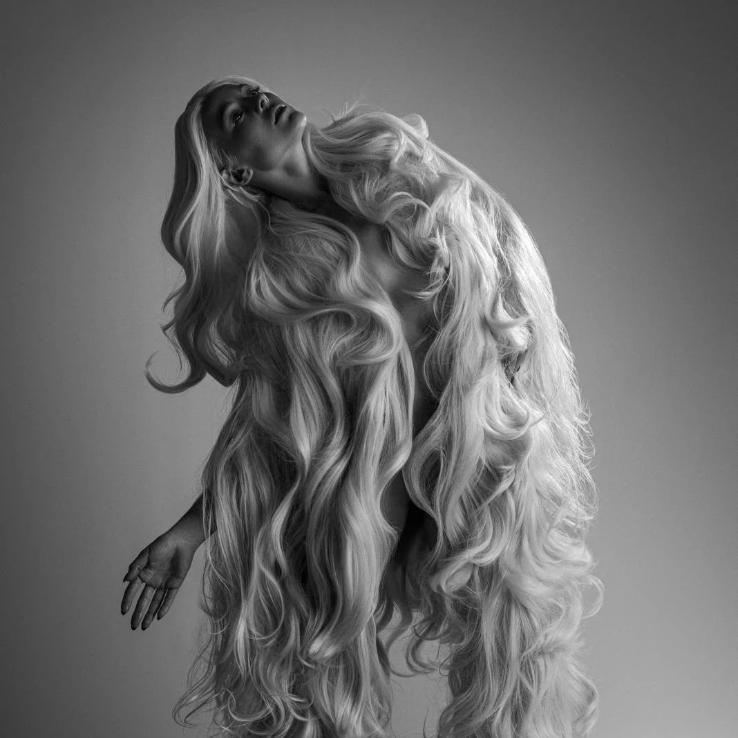 A woman with long, flowing blonde hair leaning back against a plain background in a black-and-white photo