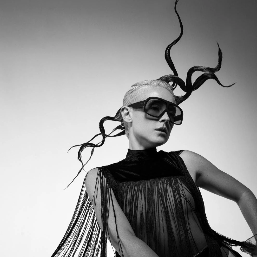 A black and white photo of a woman with platinum blonde hair, wearing large goggles, a sleeveless high-neck dress with fringe detailing, and abstract plant-like hair extensions.