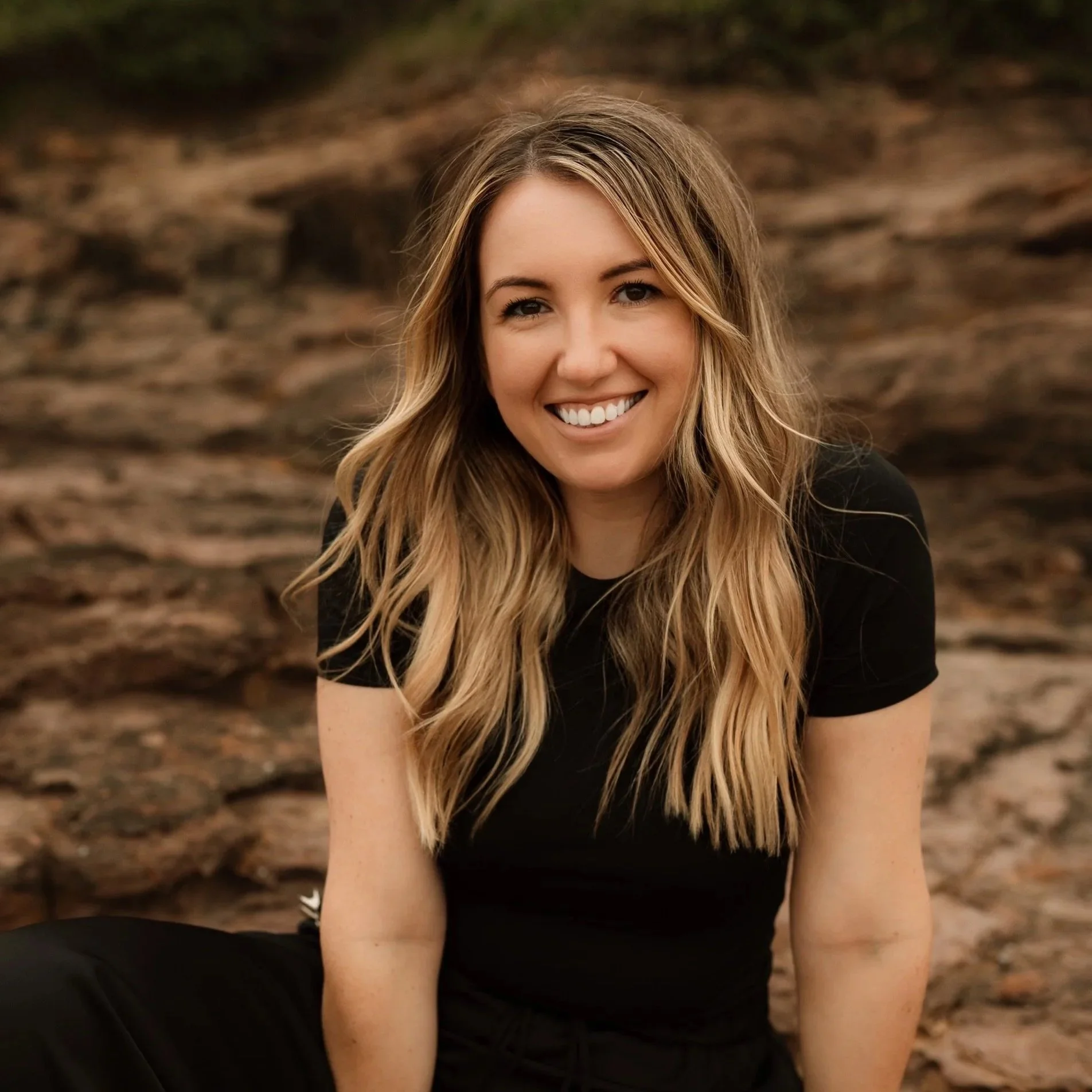A smiling woman with long wavy blond hair wearing a black shirt, sitting outdoors on a rocky terrain.