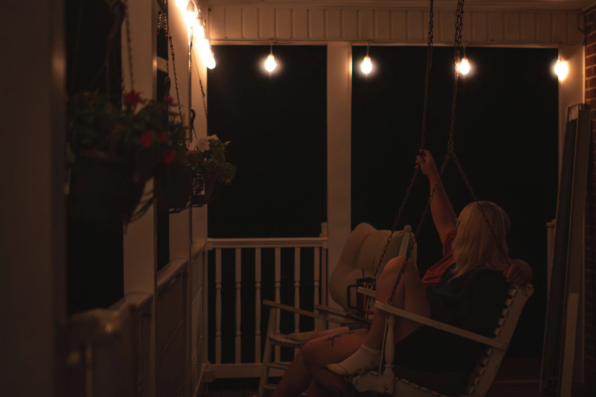 A woman with blonde hair sitting on a porch swing at night, holding onto chains, illuminated by string lights overhead.