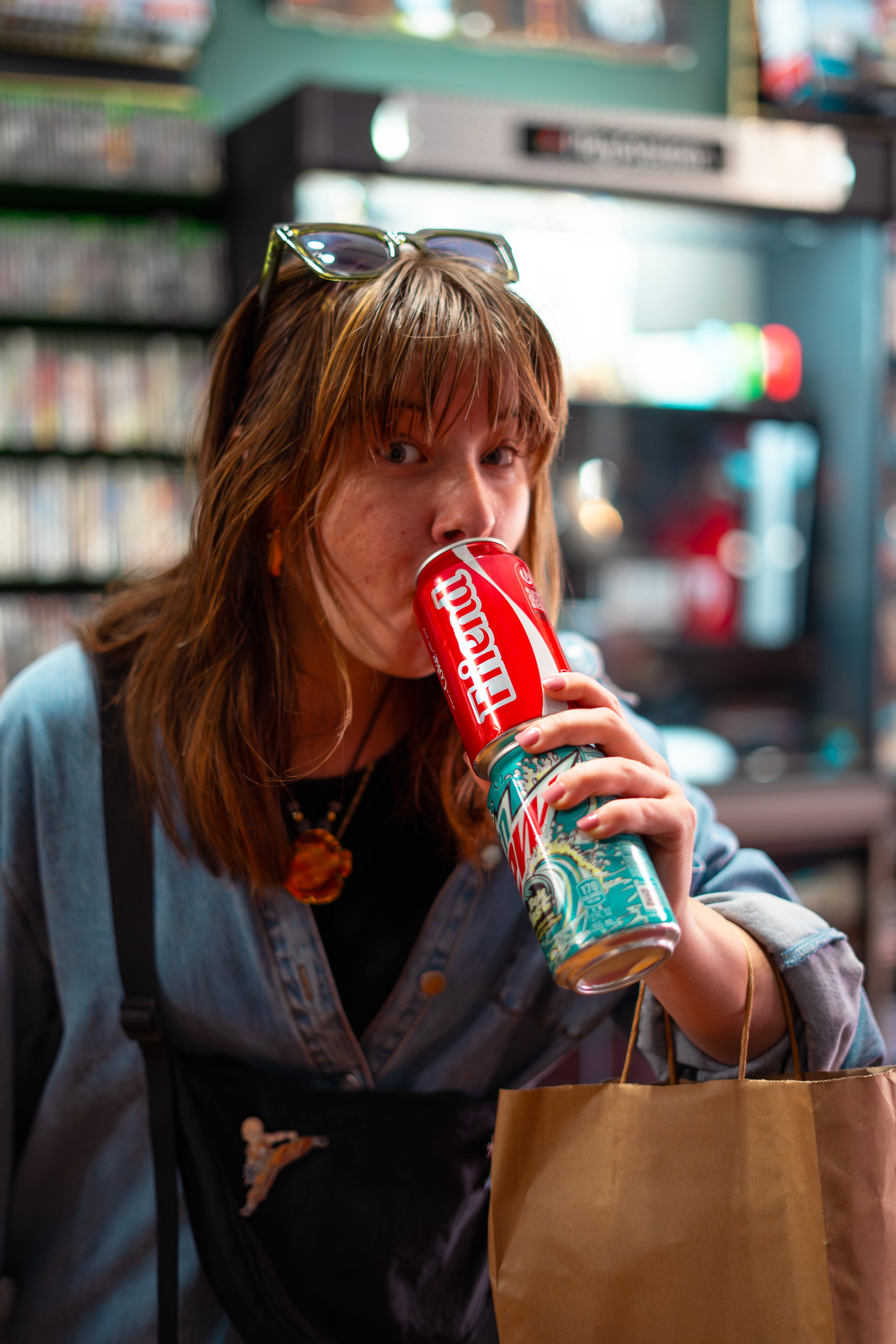A young woman with brown hair and glasses on her head drinks from a colorful can while holding a paper bag, indoors with shelves and a heater in the background.
