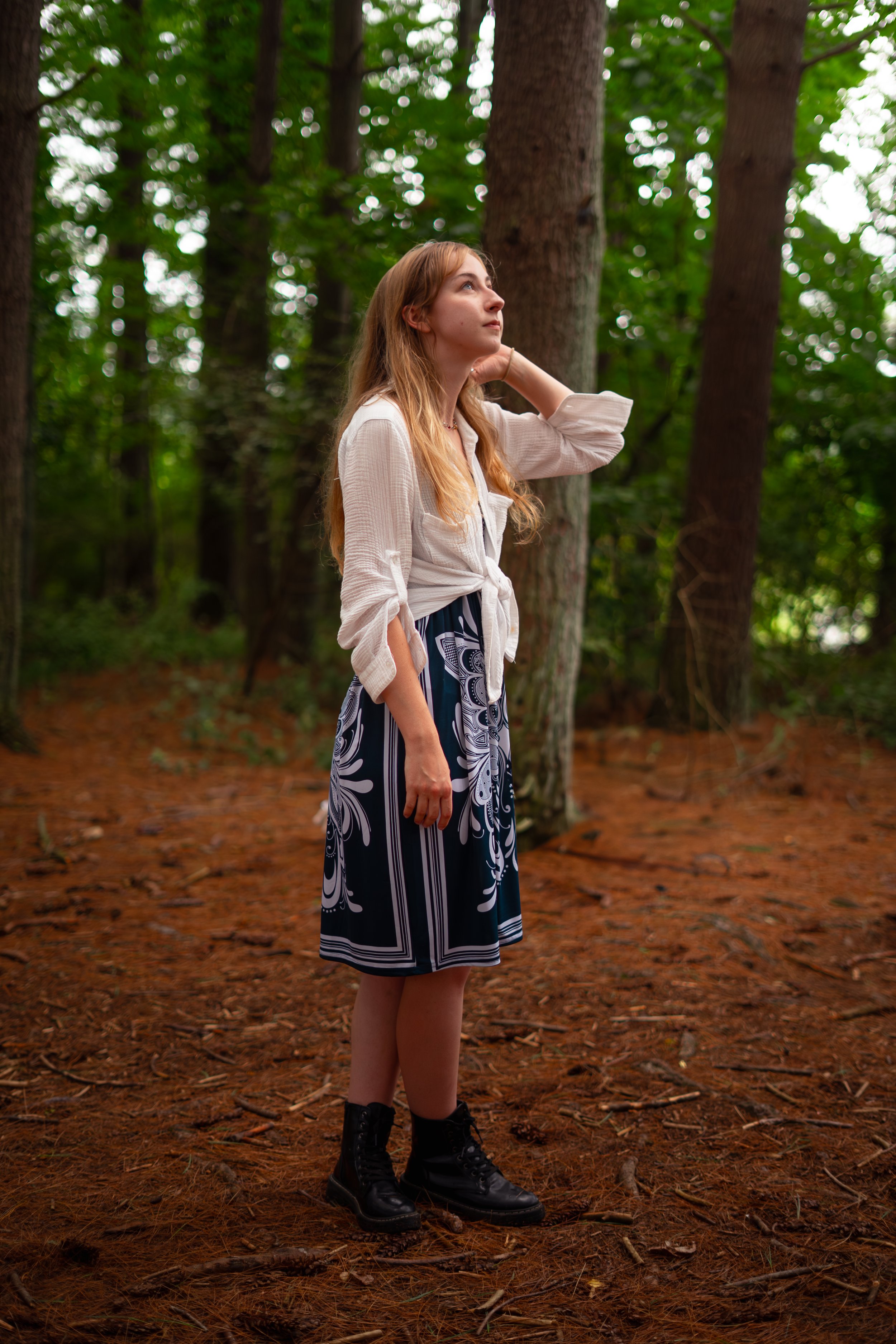 A young woman with long blonde hair, wearing a white shirt, patterned skirt, and black boots, stands in a forest with tall trees and green foliage, looking contemplatively to the side.