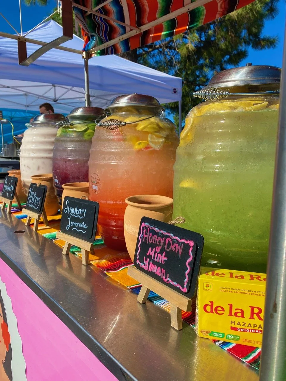 Large jars of colorful fruit-infused water at an outdoor market stand, with chalkboard signs labeling flavors like Honey Dew Mint and Strawberry Lemonade, under a vibrant striped canopy and white tent. Chuliutas Modern Mexican Food. Century Commons.