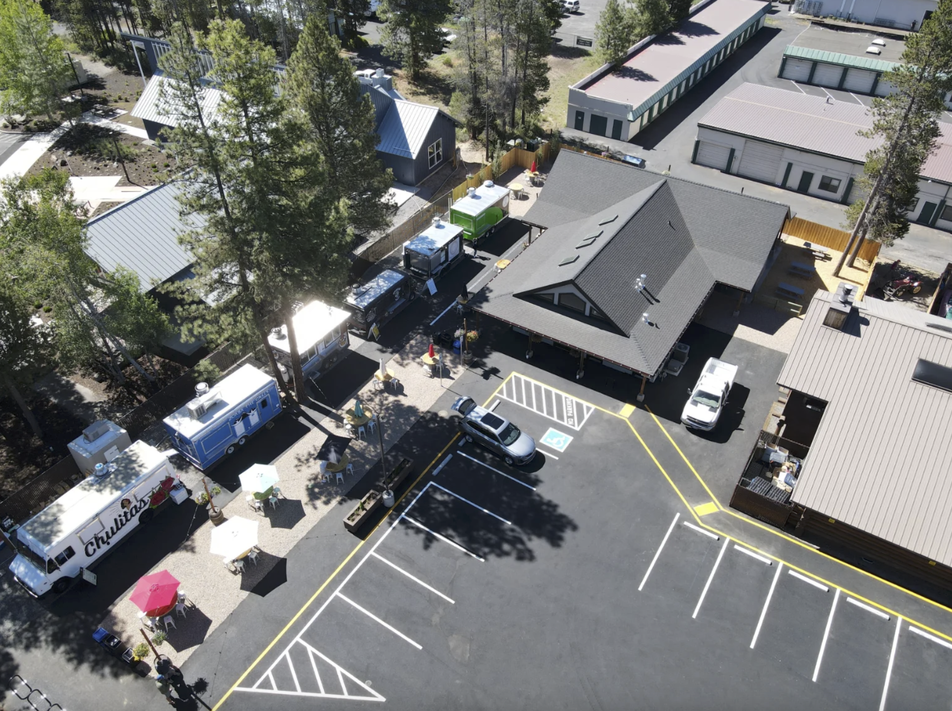 Aerial view of Century Commons Taps & Trucks with food trucks, an outdoor seating area with tables and umbrellas, surrounded by trees and the tap house, including a parking lot in the foreground. Shows the large parking lot and size of tap house.