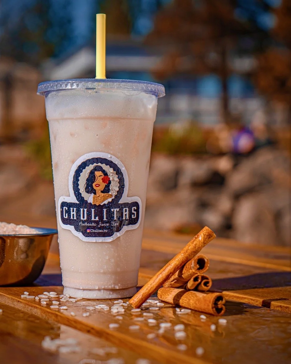A clear plastic cup with a straw and a lid, labeled 'Chulitas Authentic Juice Bar,' containing a creamy, frothy beverage on a wooden table. Next to the cup are cinnamon sticks and white beans or sugar crystals, Chulitas Modern Mexican Food