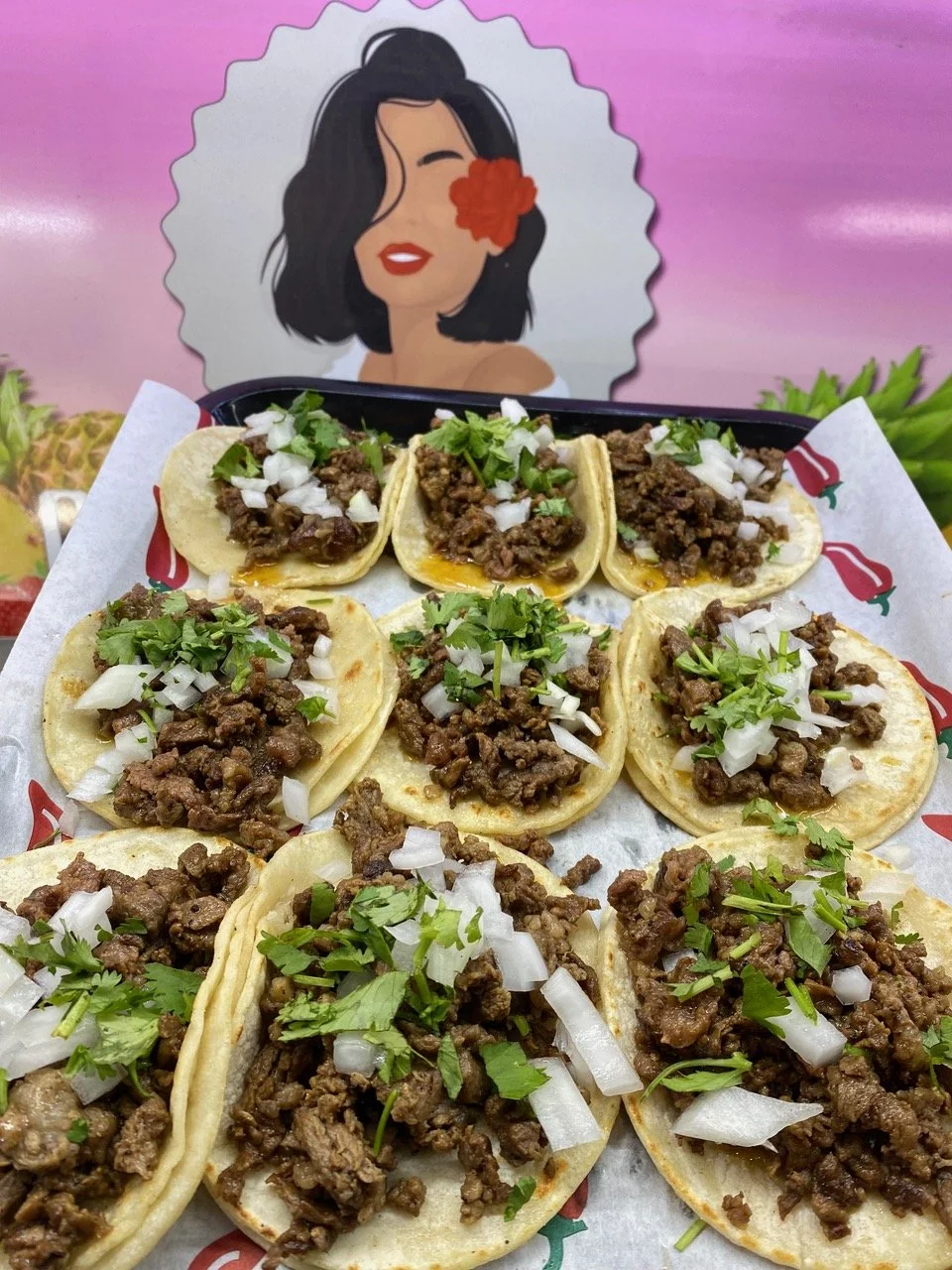 A tray of street tacos filled with seasoned ground beef, topped with chopped onions and cilantro, served on small tortillas against a pink background with a cartoon woman illustration. Central Oregon, Sunriver food trucks, Century Commons Taps.