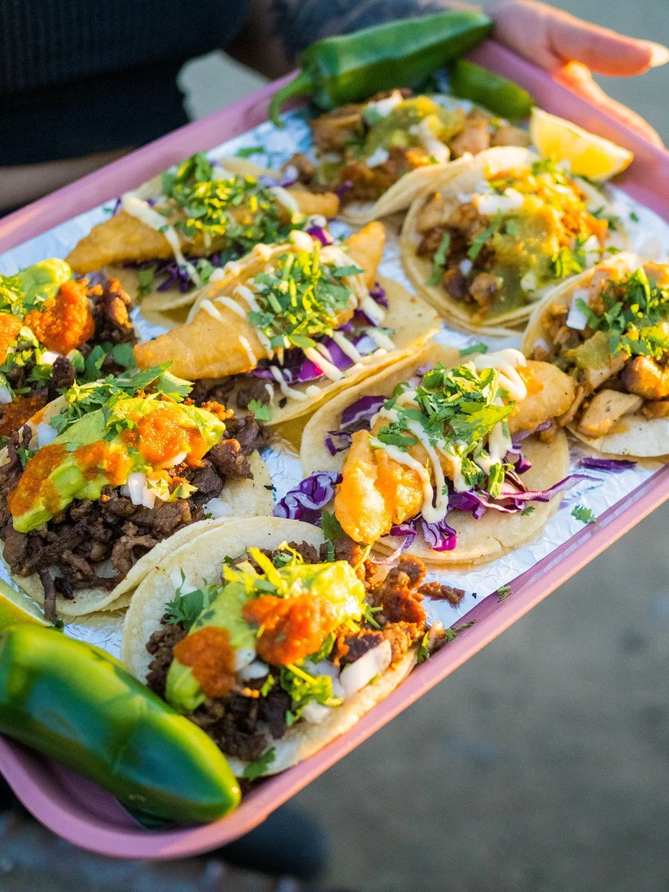 A pink tray with multiple small tacos filled with meat, fish, and pork, topped with chopped cilantro, and sauces, garnished with lime wedges, salsa, avocado, and jalapeños. Central Oregon, Sunriver food trucks, Century Commons Taps and Trucks.