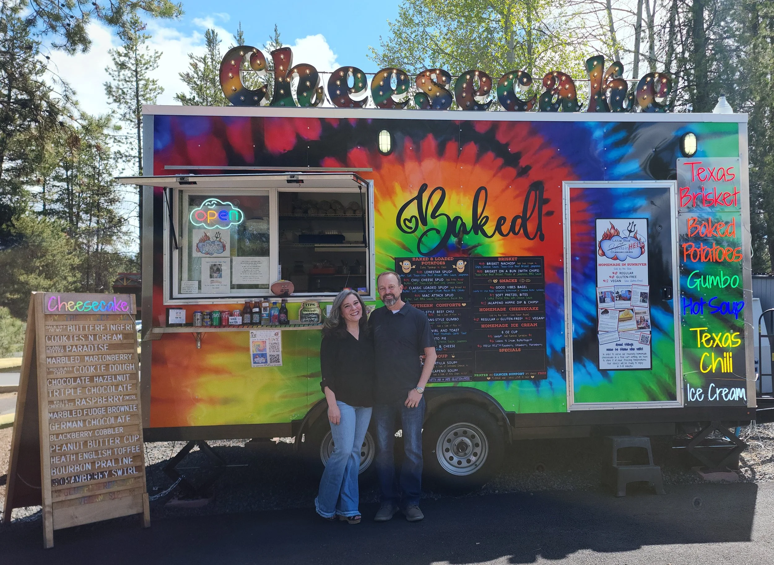 A colorful food truck named "Cheesecake" with a rainbow tie-dye background, serving baked goods like cheesecake and various flavors of ice cream. Two people are standing in front of the truck, smiling. The truck menu lists items such as Texas Brisket