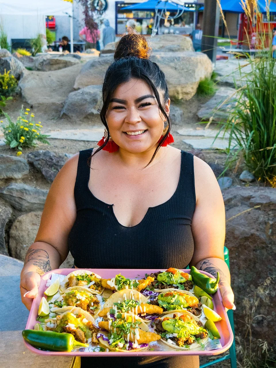 A smiling woman holding a pink tray of tacos, burritos, and quesadillas outdoors with rocks and plants in the background. Chulitas Modern Mexican Food. Central Oregon, Sunriver food trucks, Century Commons Taps and Trucks. 