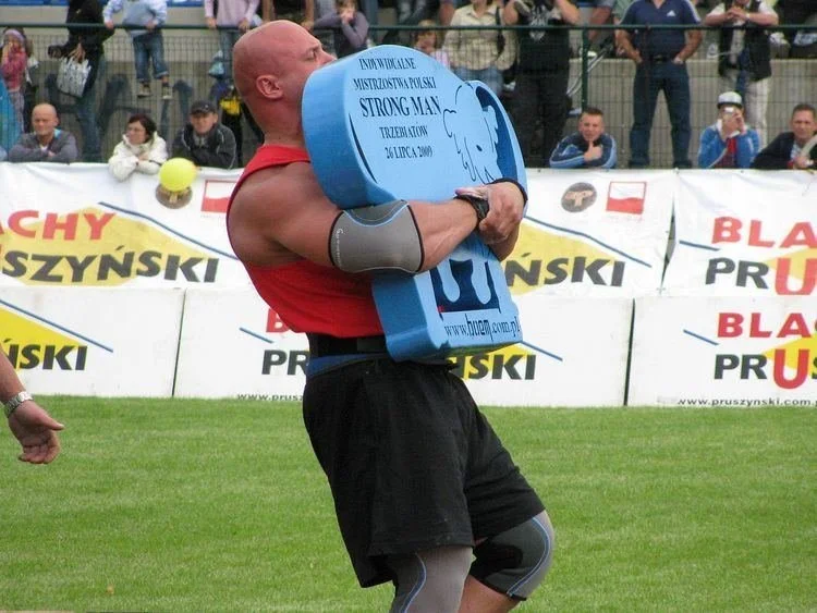 A strongman athlete in a front-carry position with a black Husafell sandbag, demonstrating the classic Icelandic stone lift technique during a heavy workout.
