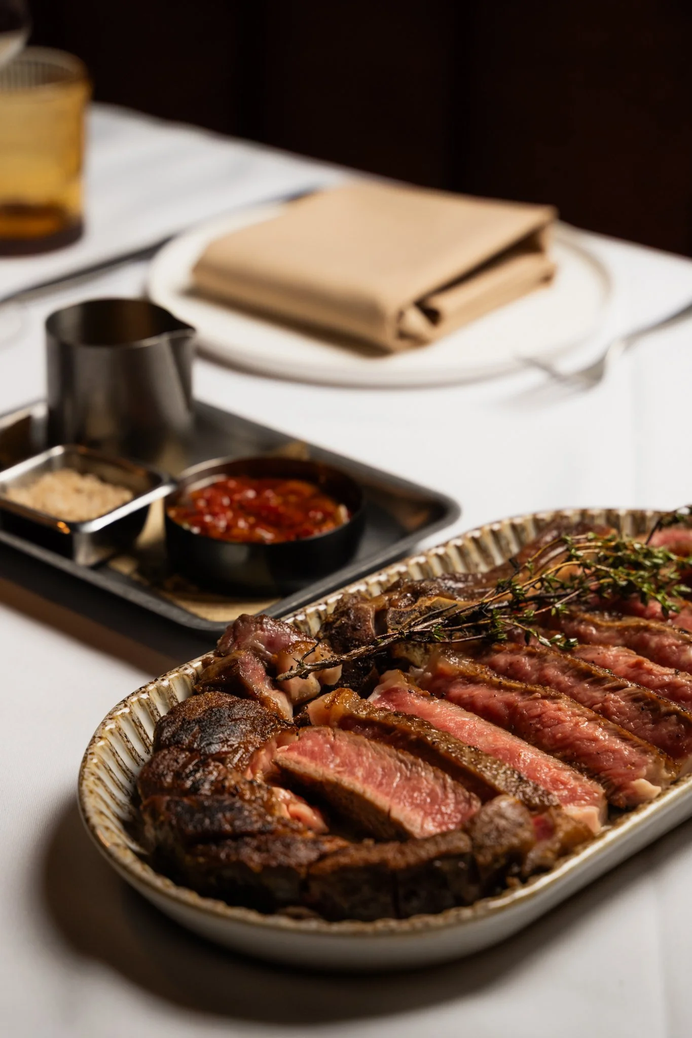 A platter of sliced medium-rare steak on a table with condiments and utensils, restaurant setting.