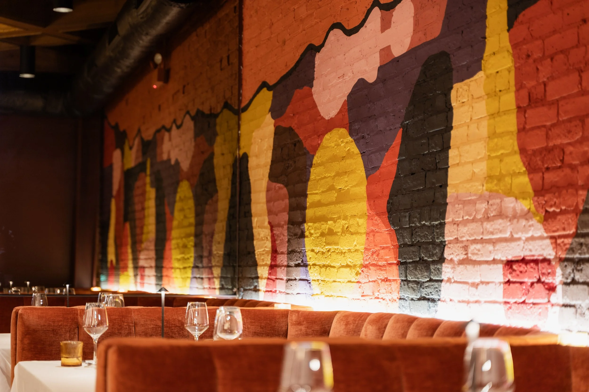Interior of a restaurant with a brick wall painted with colorful abstract art, orange upholstered chairs, and tables set with wine glasses and small candles.