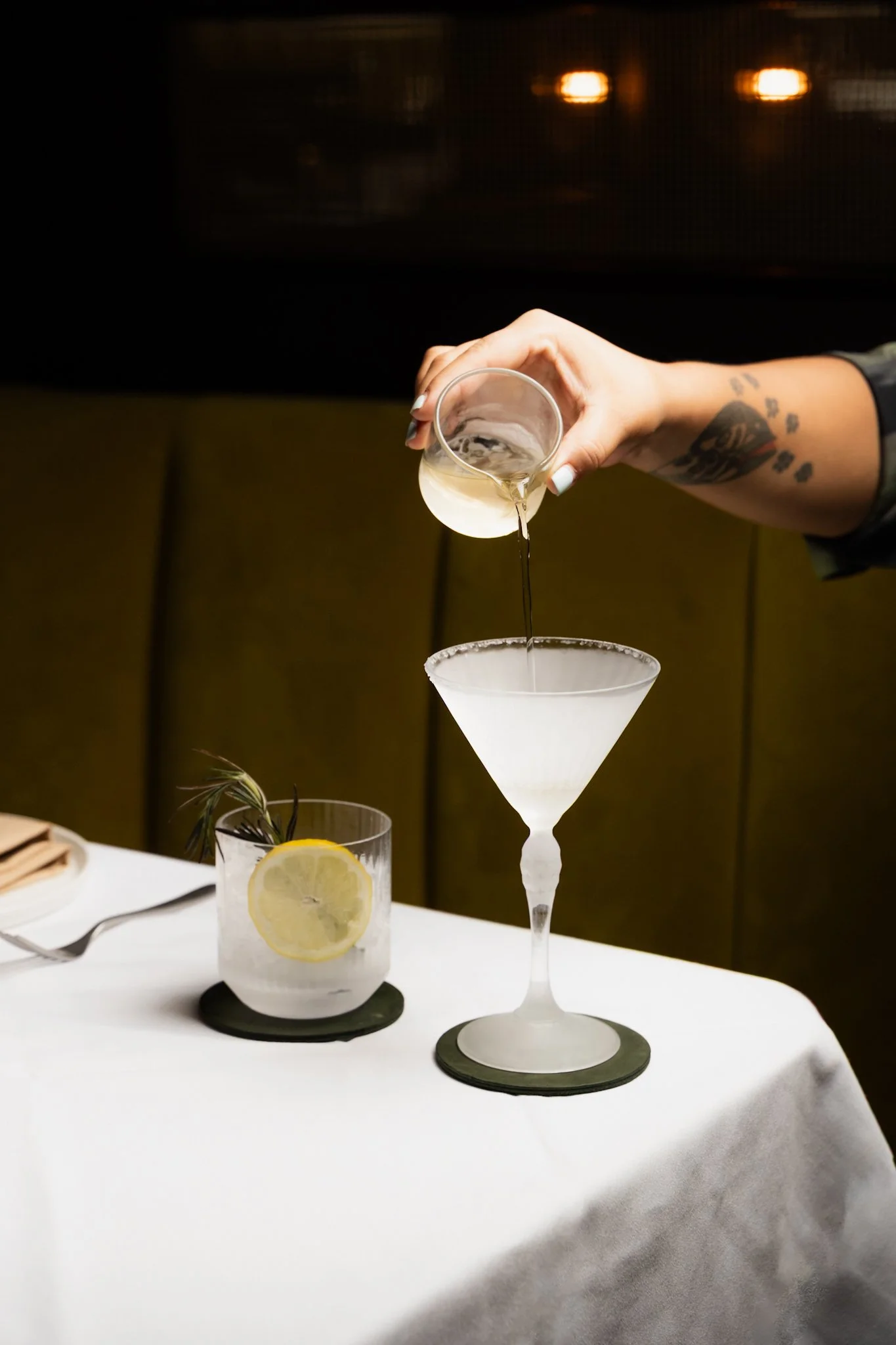A person pours a clear liquid into a white cocktail glass on a table, with a glass of water with lemon and a sprig of rosemary nearby.