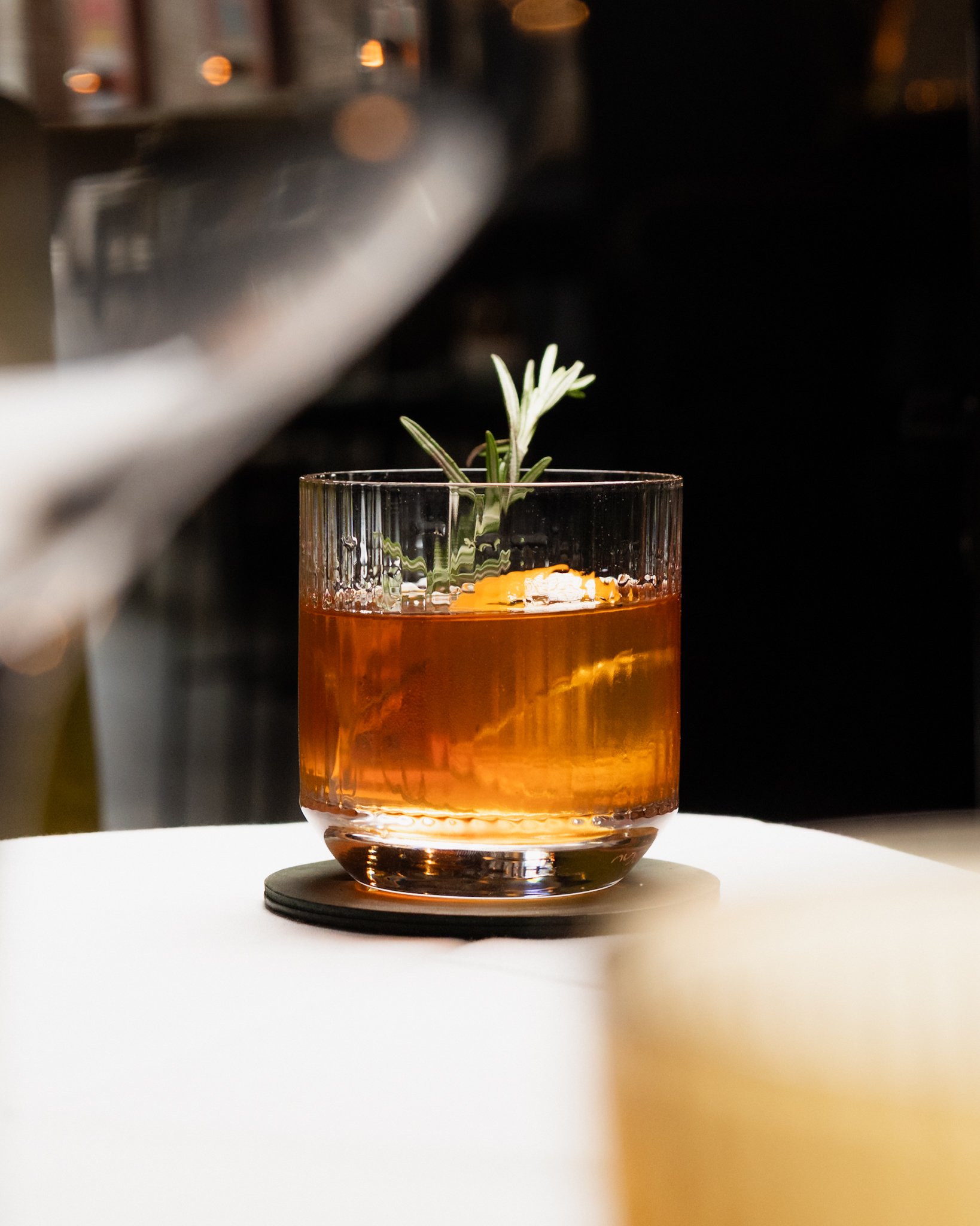 A glass of amber-colored cocktail with a sprig of rosemary garnish on a white surface.