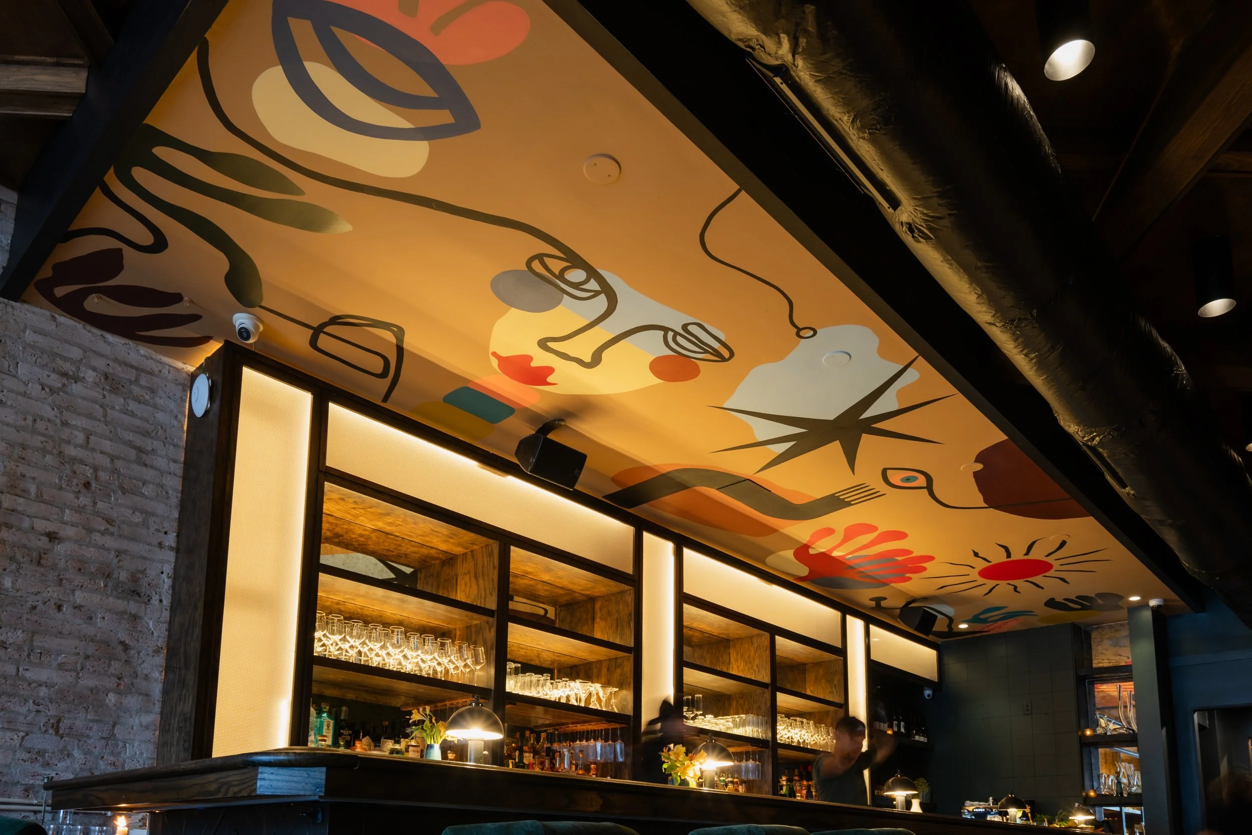 Interior of a restaurant bar with a colorful abstract pattern on the ceiling, a wooden bar counter with glasses, and shelves with bottles and glasses in the background.