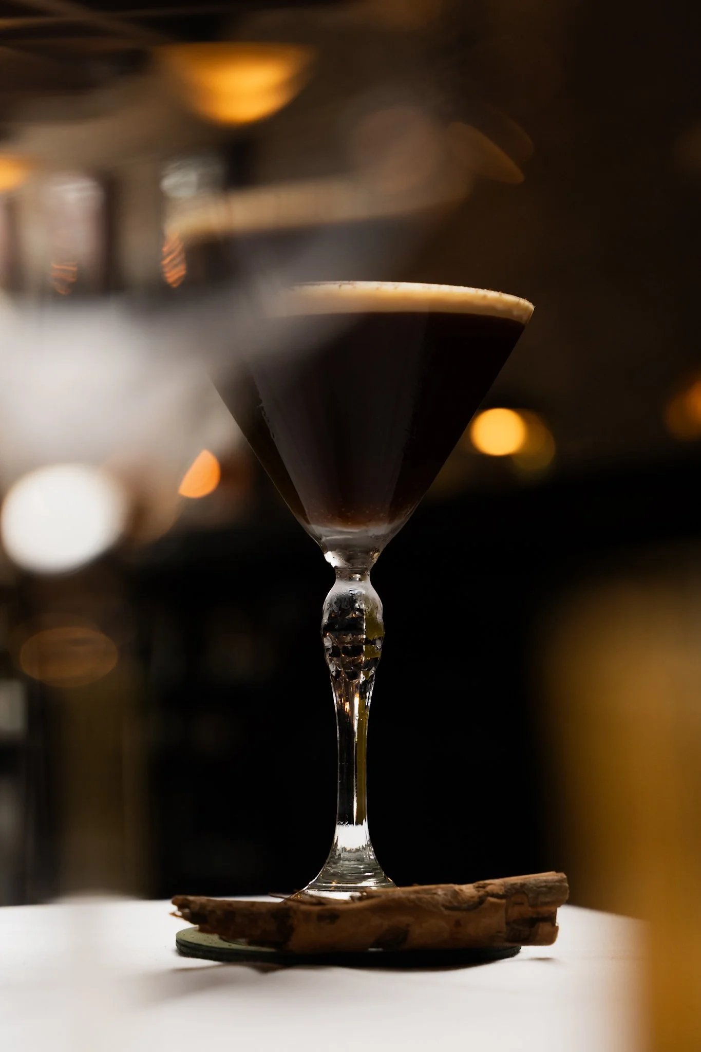 A martini glass filled with a dark cocktail, garnished with a lemon twist, placed on a piece of driftwood, with blurred warm lights in the background.