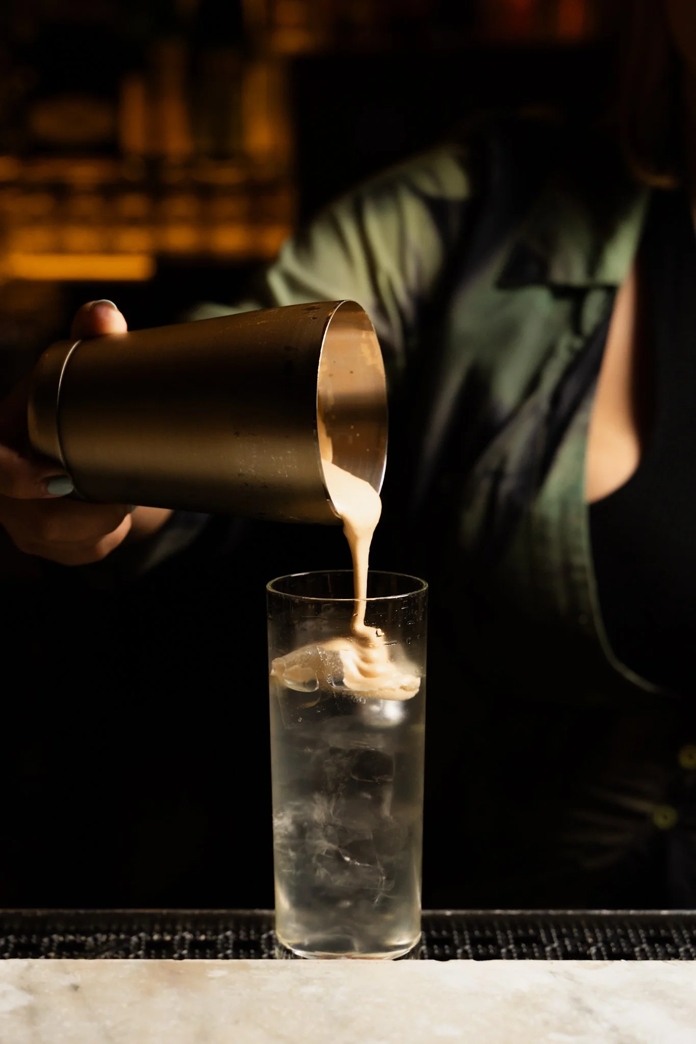 A person pouring a creamy cocktail from a cocktail shaker into a tall glass with ice cubes and lemon slices, against a dark background.