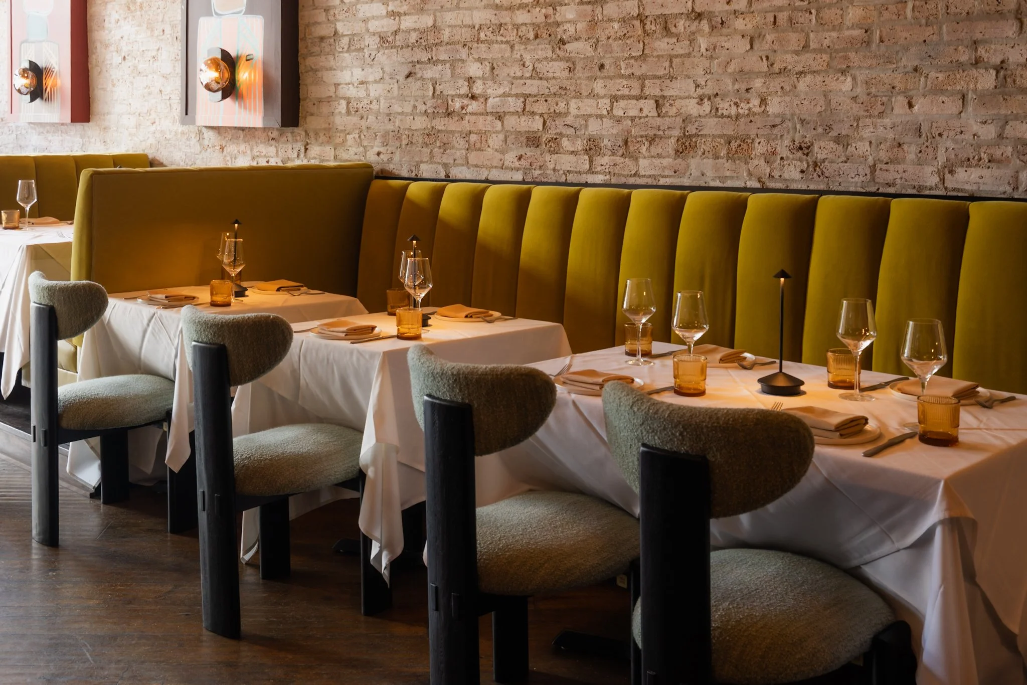 Upscale restaurant dining table set with white tablecloths, wine glasses, amber glassware, beige napkins, and modern black lamps. Green velvet banquette seating along a brick wall, with upholstered chairs in the foreground.