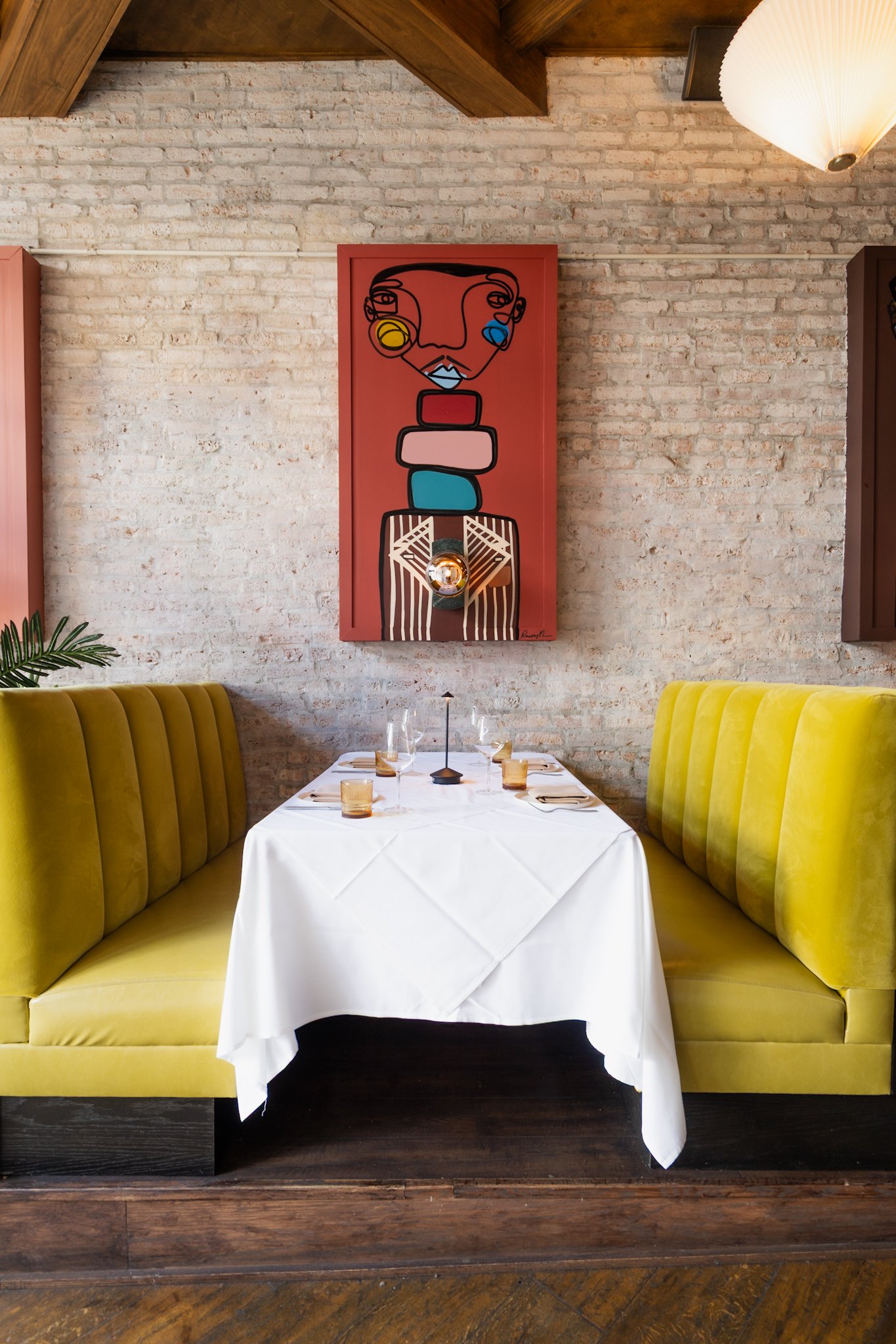 A dining table set with a white tablecloth, glasses, and utensils, surrounded by yellow upholstered booths, with a brick wall background in a restaurant, featuring a colorful abstract artwork of a face on the wall.