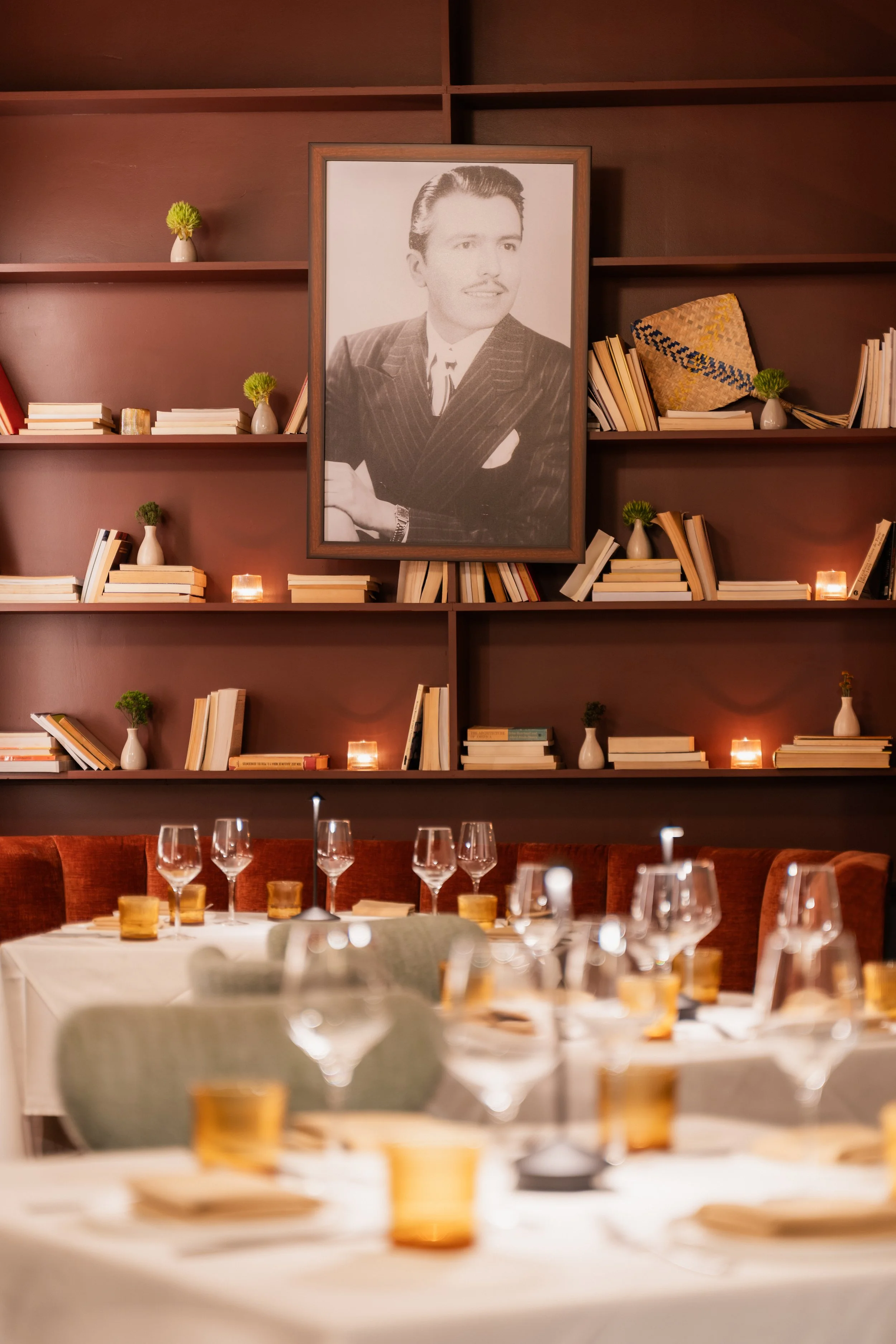 Elegant restaurant interior with neatly arranged tables, glasses, and candles, featuring a large framed black-and-white portrait of a man in a suit on a bookshelf wall decorated with books and small vases.