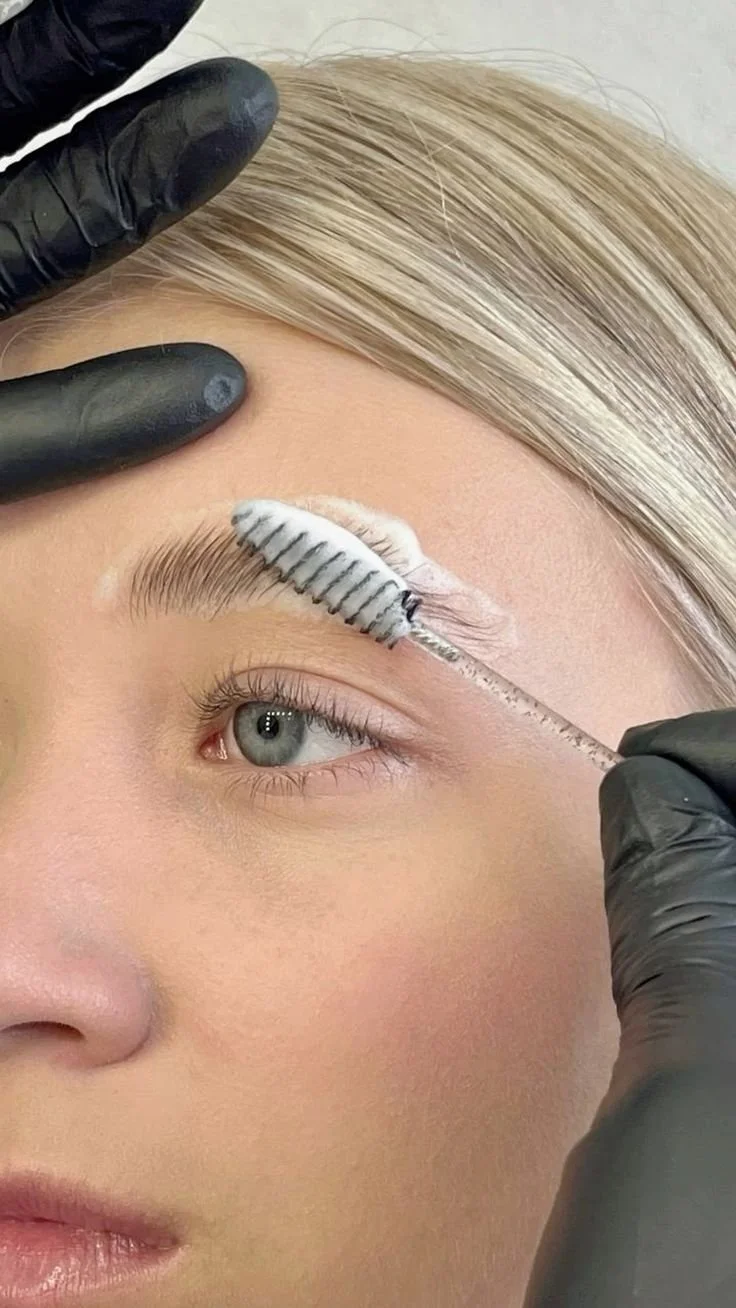 A person receiving an eyelash extension treatment, with a closeup of their eye and eyebrow. A gloved hand is applying the treatment with a tool that has a white and black-striped extension on it.