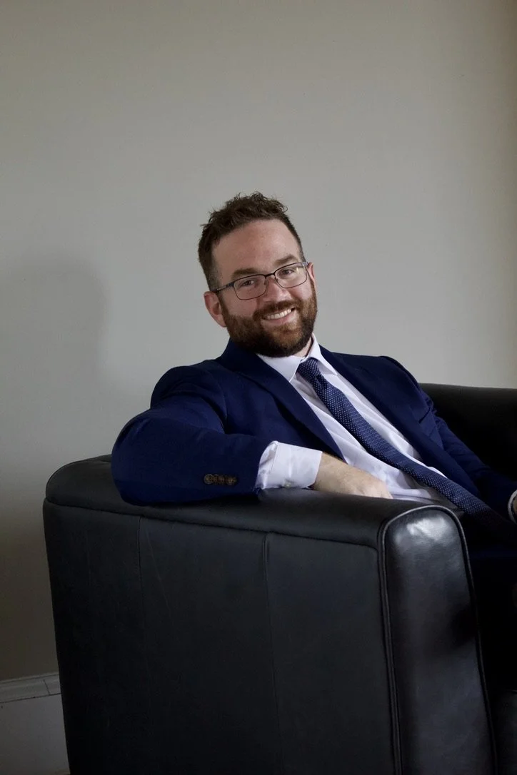 A man (Attorney Tyler Crawford of Lighthouse Legal) with glasses, a beard, and brown hair, wearing a navy blue suit, white shirt, and tie, sitting on a black sofa against a plain wall, smiling at the camera.