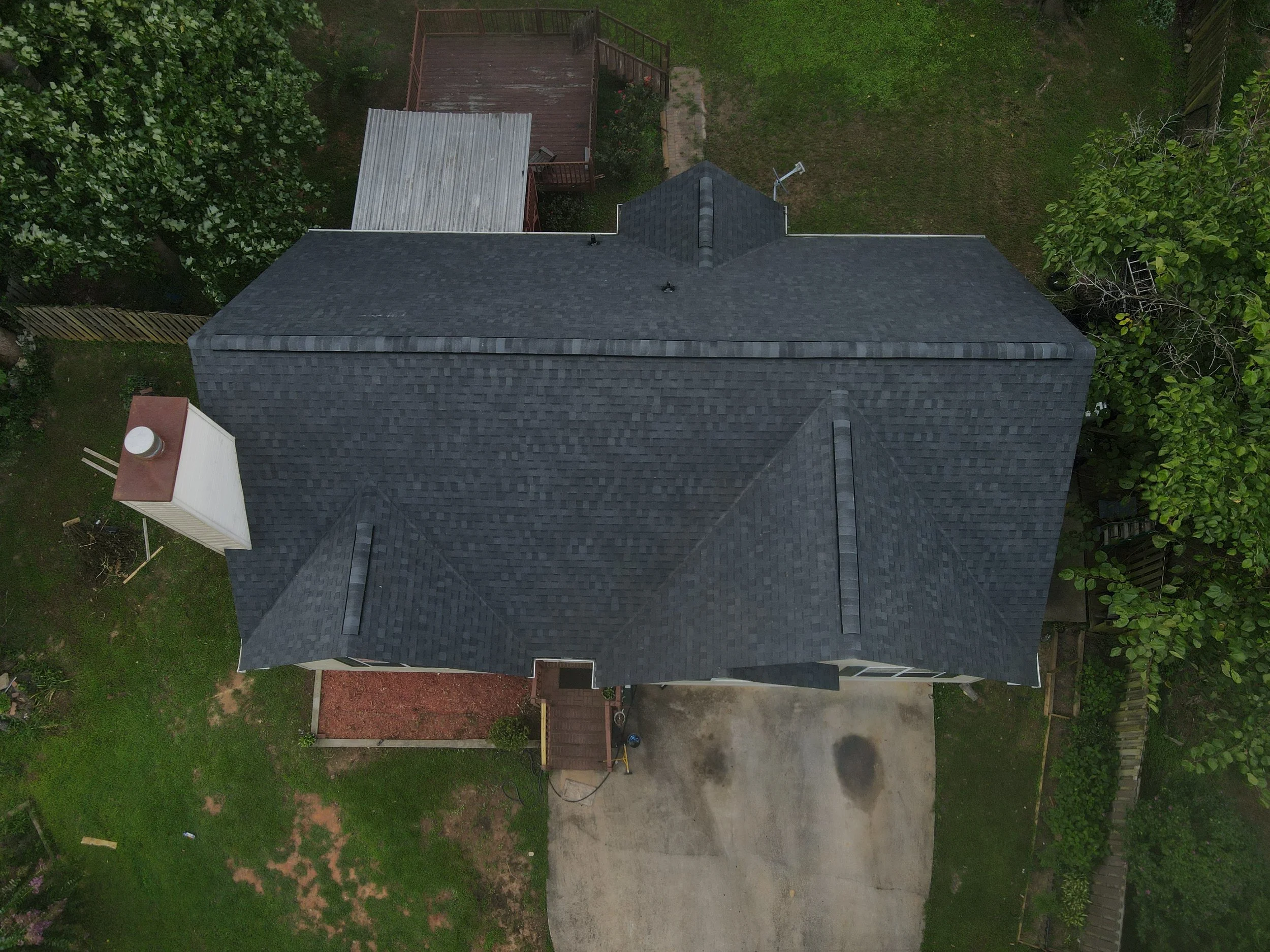 Aerial view of a house with dark gray roof shingles, a driveway, and surrounding green yard with trees and a fenced backyard.