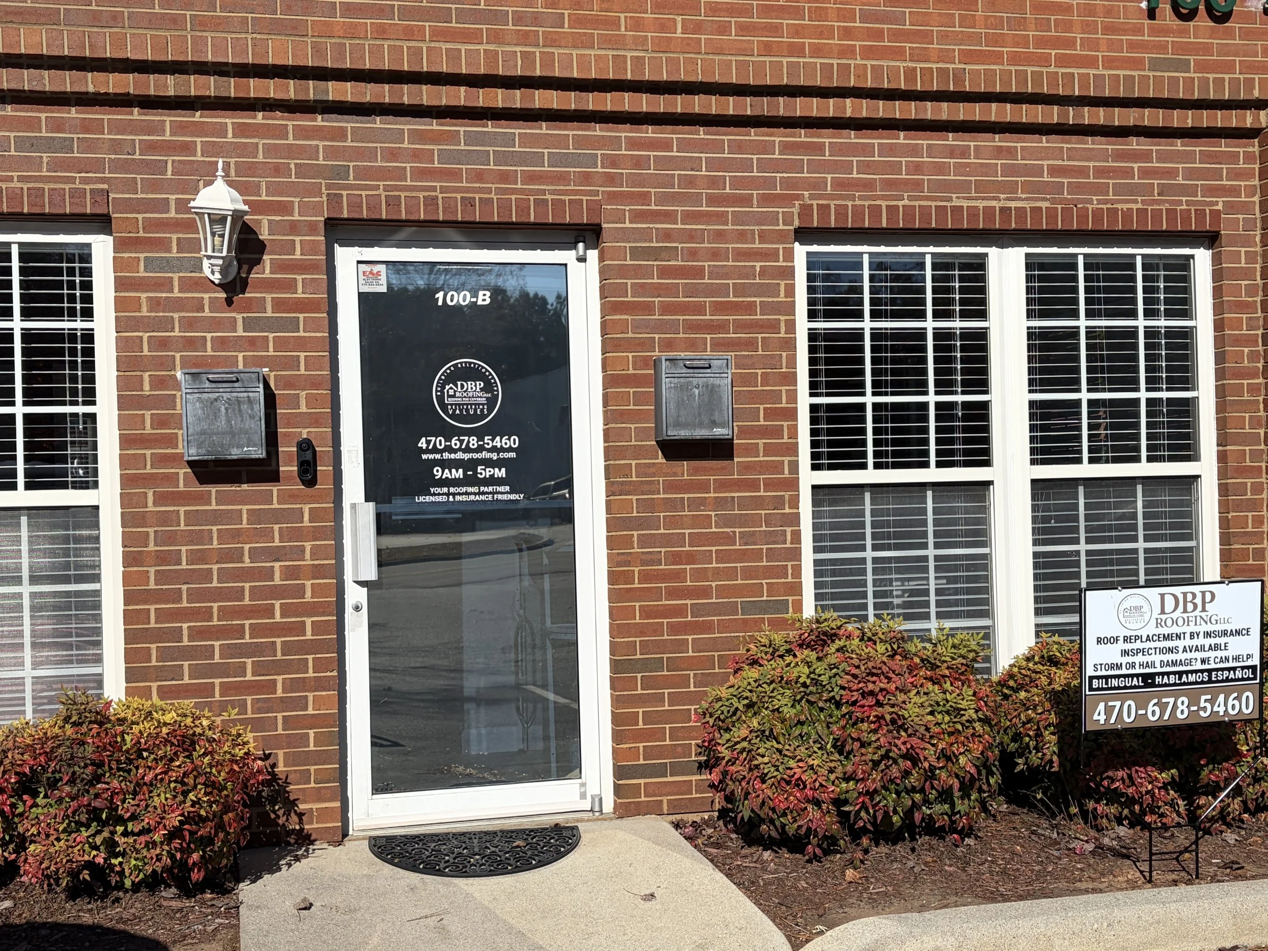Brick building with a glass door, two large windows with blinds, a white outdoor light fixture, mailboxes, and a sign for a roofing company.