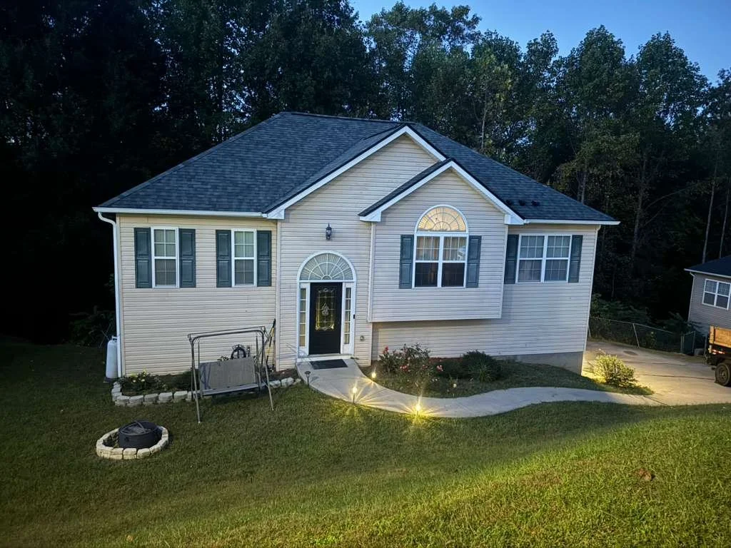 A two-story house with beige siding, blue shutters, and a dark roof. The front yard has a curved walkway, small garden, and outdoor swing, illuminated by landscape lights. There are trees in the background.