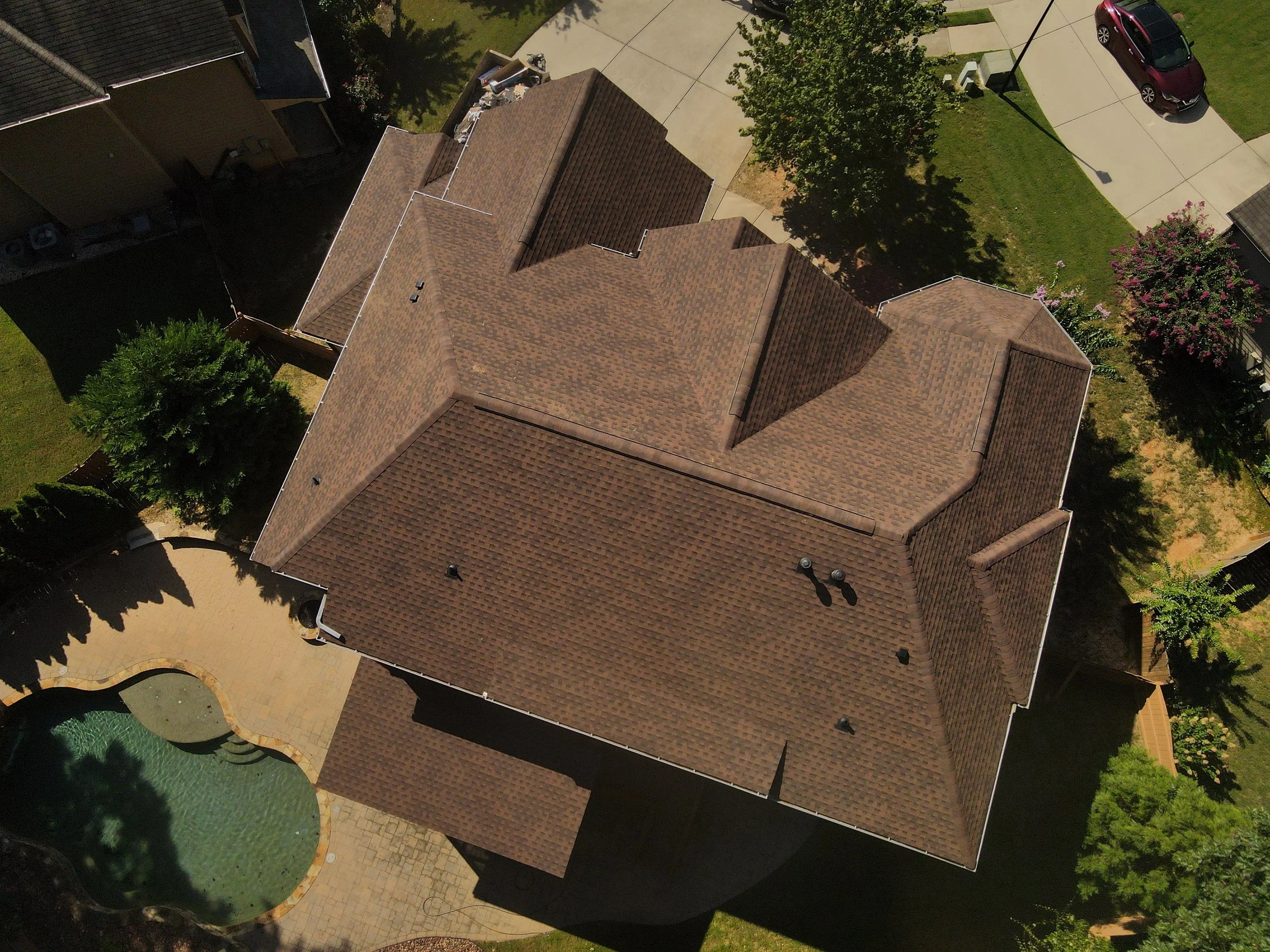  aerial view of a house with a brown shingle roof, surrounded by trees, a driveway, and a backyard with a swimming pool.