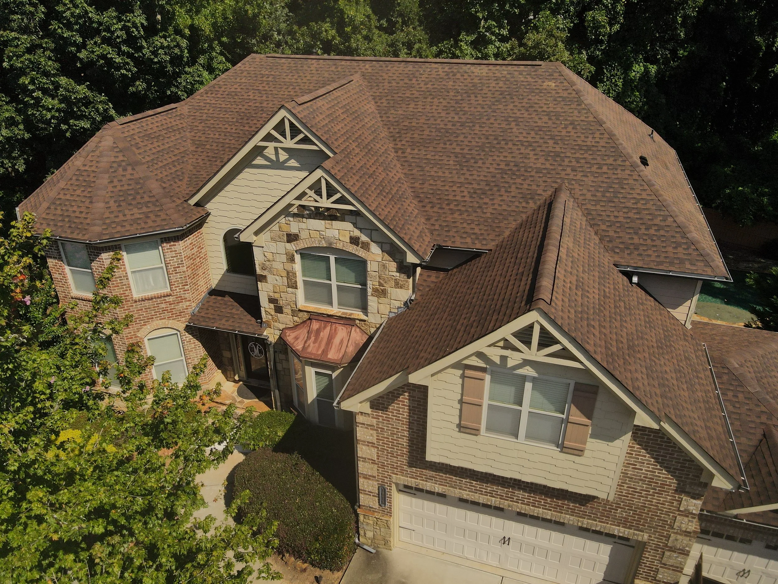 Aerial view of a large suburban house with multiple roof sections, brick and stone exterior, and a two-car garage. Trees surround the property.