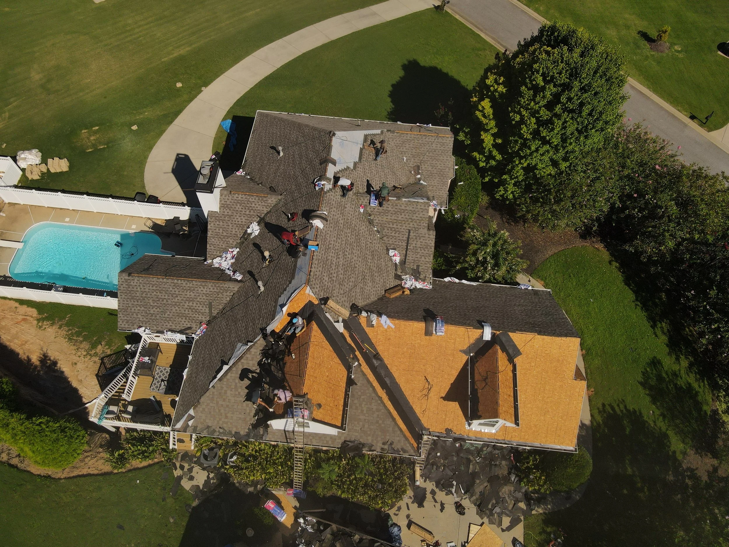Aerial view of a house under construction or renovation with workers on the roof, surrounded by a backyard with a swimming pool, trees, and green lawns.