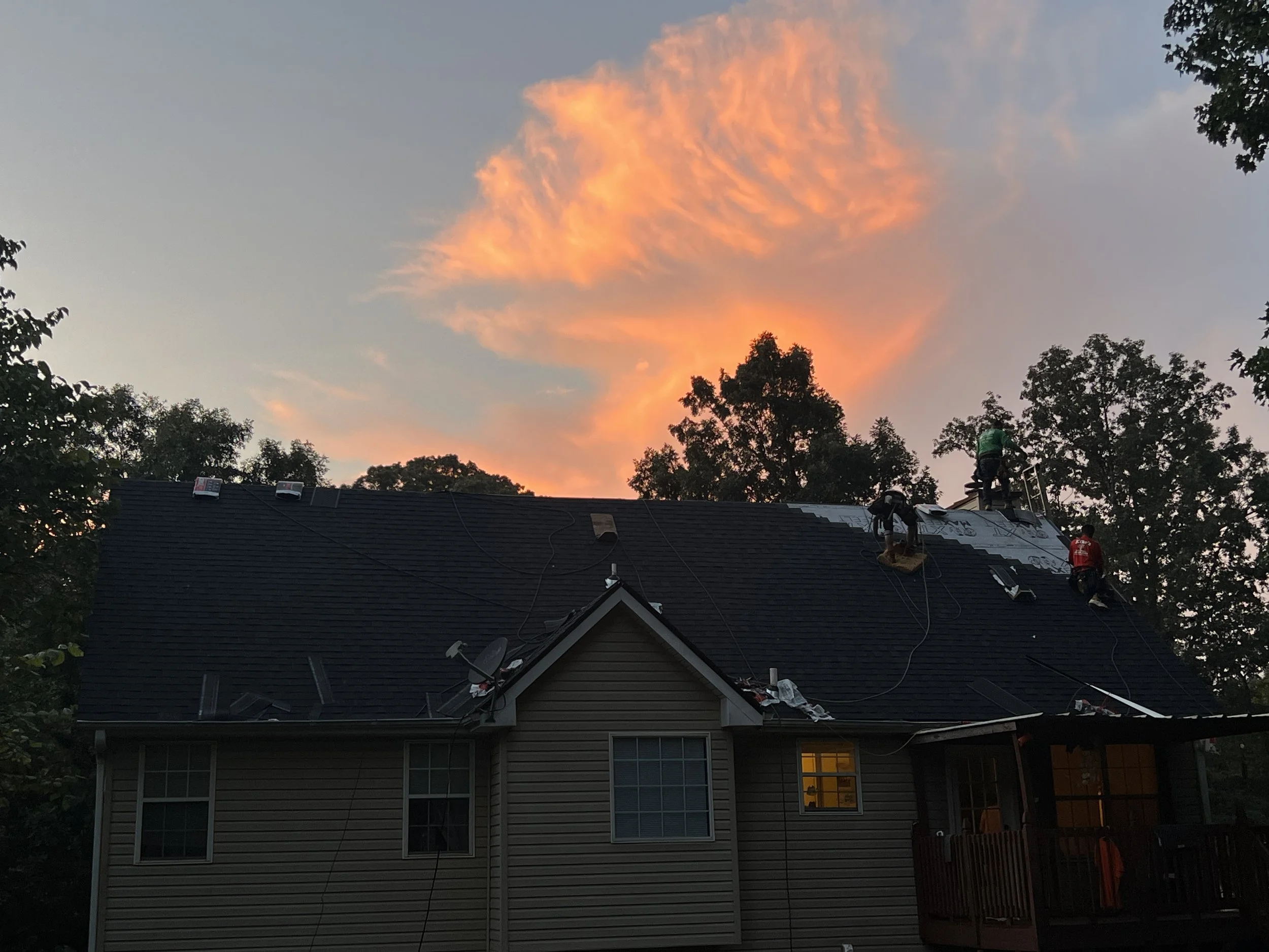 House with workers on the roof during sunset, replacing shingles, with trees and a colorful sky in the background.