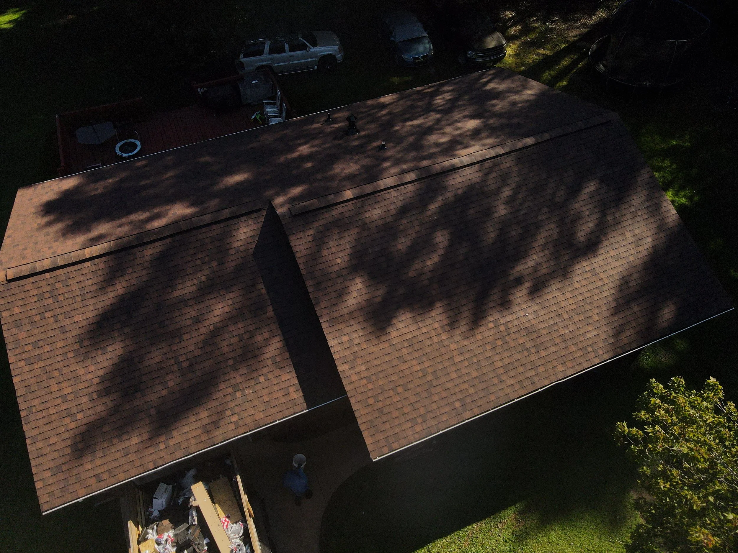 An aerial view of a house with a brown shingle roof, surrounded by trees and vehicles parked nearby.