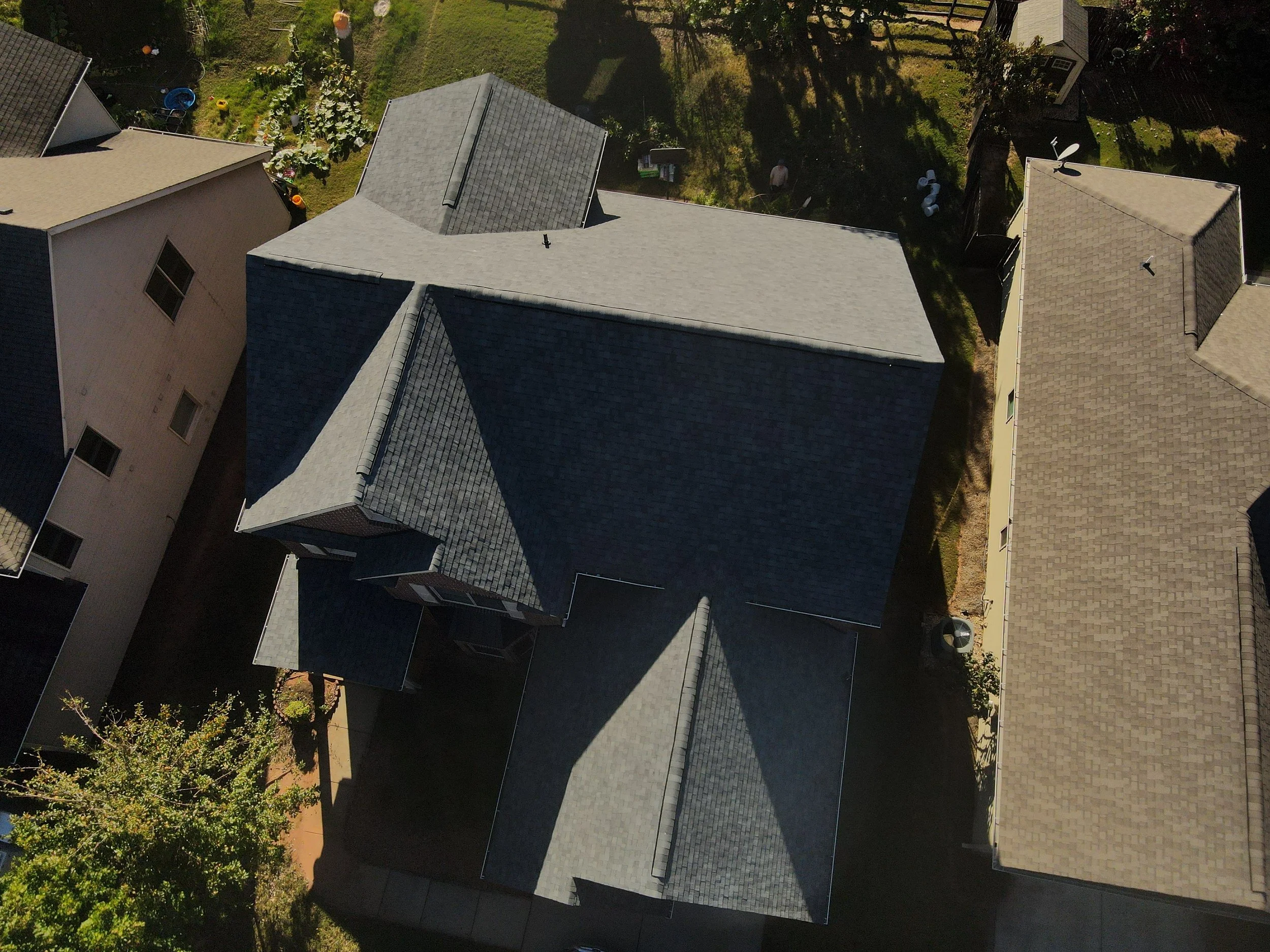 Aerial view of houses with gray and beige rooftops, a backyard with greenery, and shadows cast by trees.