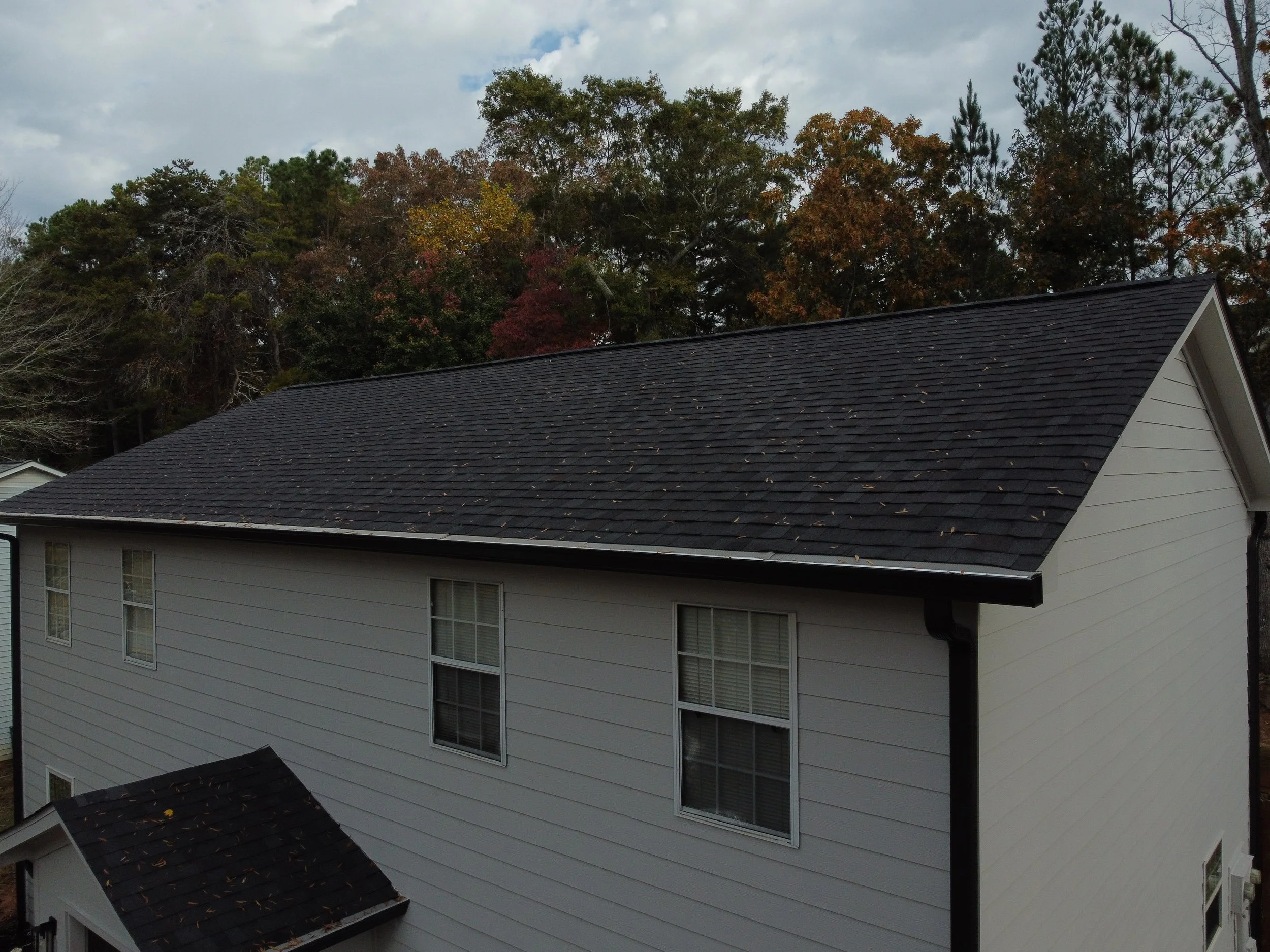 Upper view of a house with black shingle roof, white siding, and surrounding trees with autumn foliage in the background.