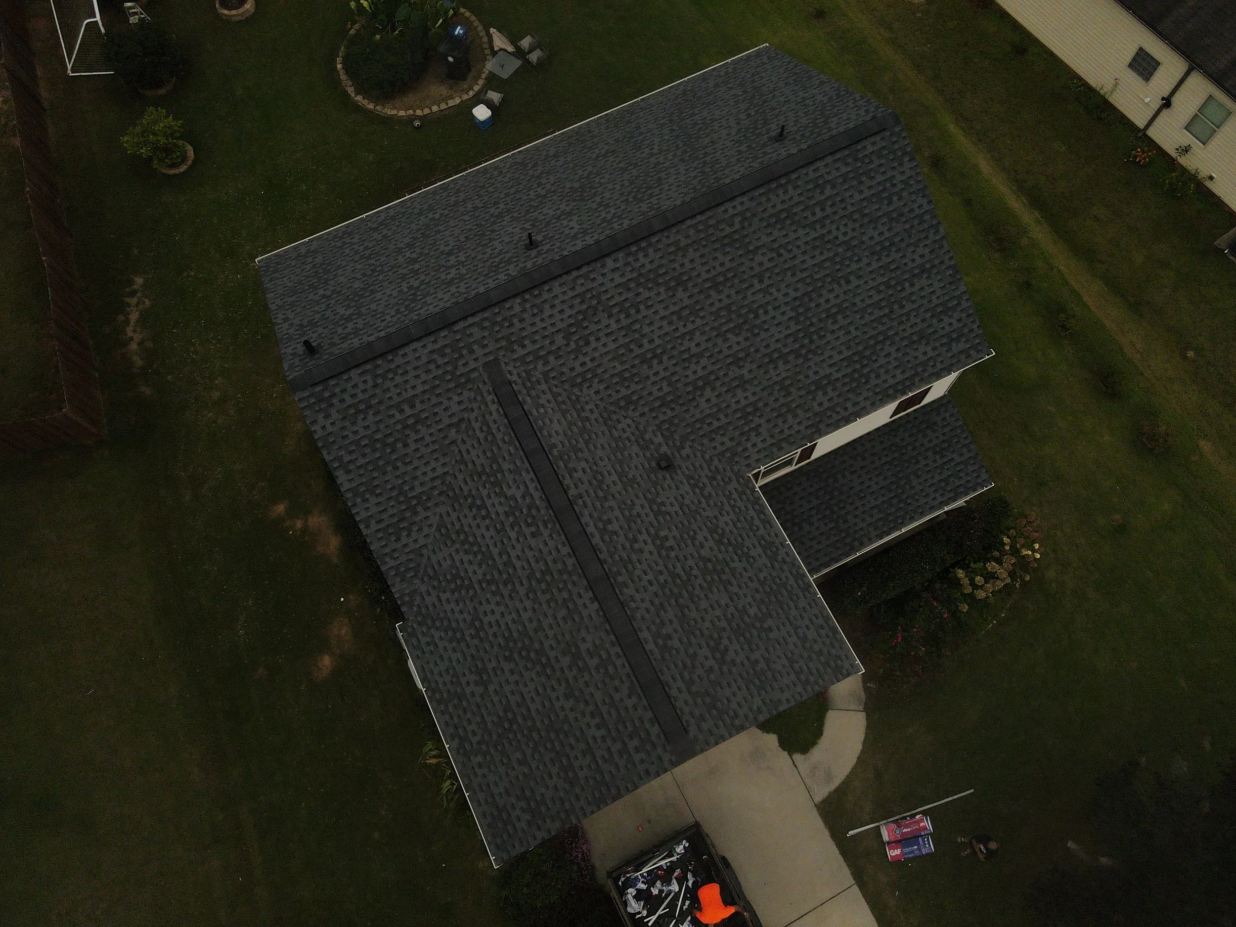 Aerial view of a house with a dark gray roof, surrounded by a green yard with a garden, a small circular patio, and some tools and equipment near the driveway.