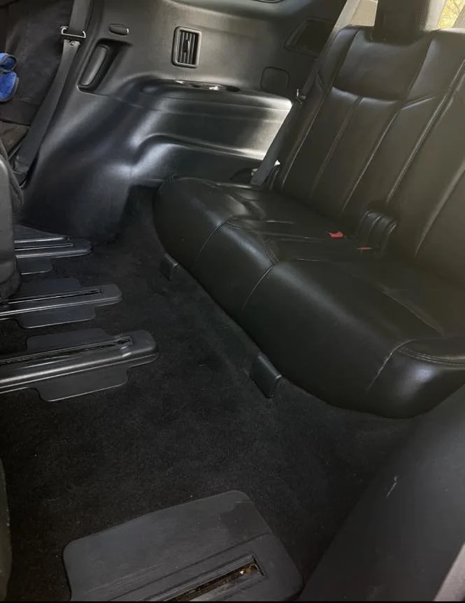 Interior of a vehicle showing the back seat with black leather upholstery and seating for three passengers. The floor is carpeted in black, and part of the front seat leg area is visible.