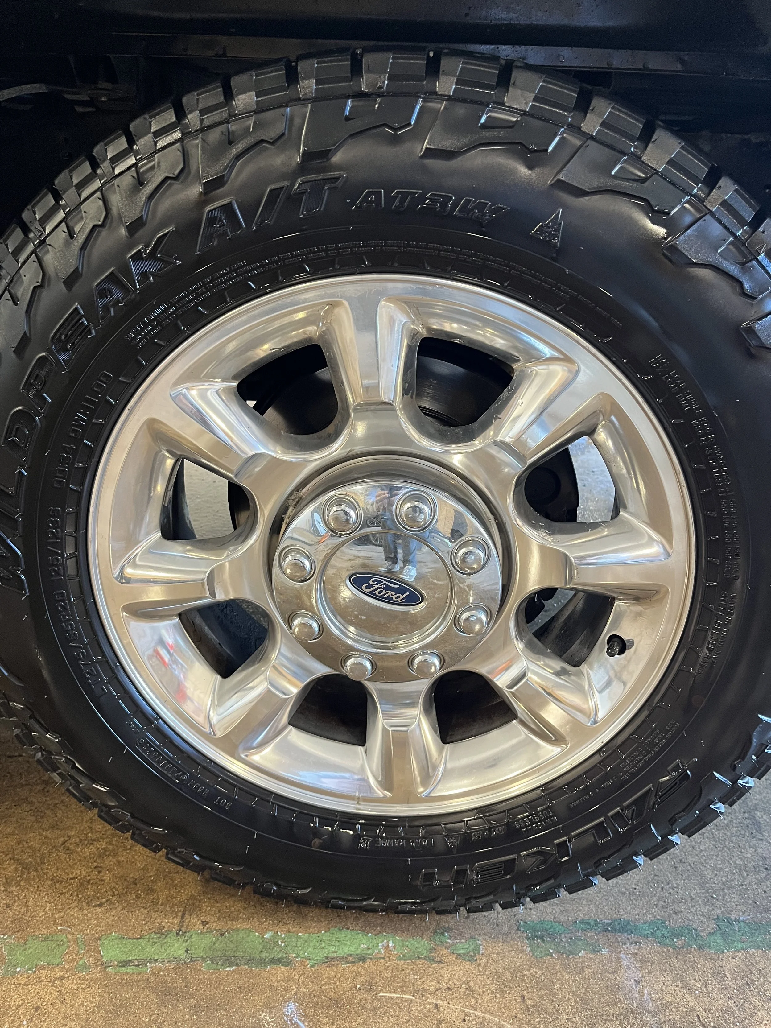 Close-up of a Ford vehicle wheel with a chrome rim and large off-road tire. The Ford logo is visible on the center cap.