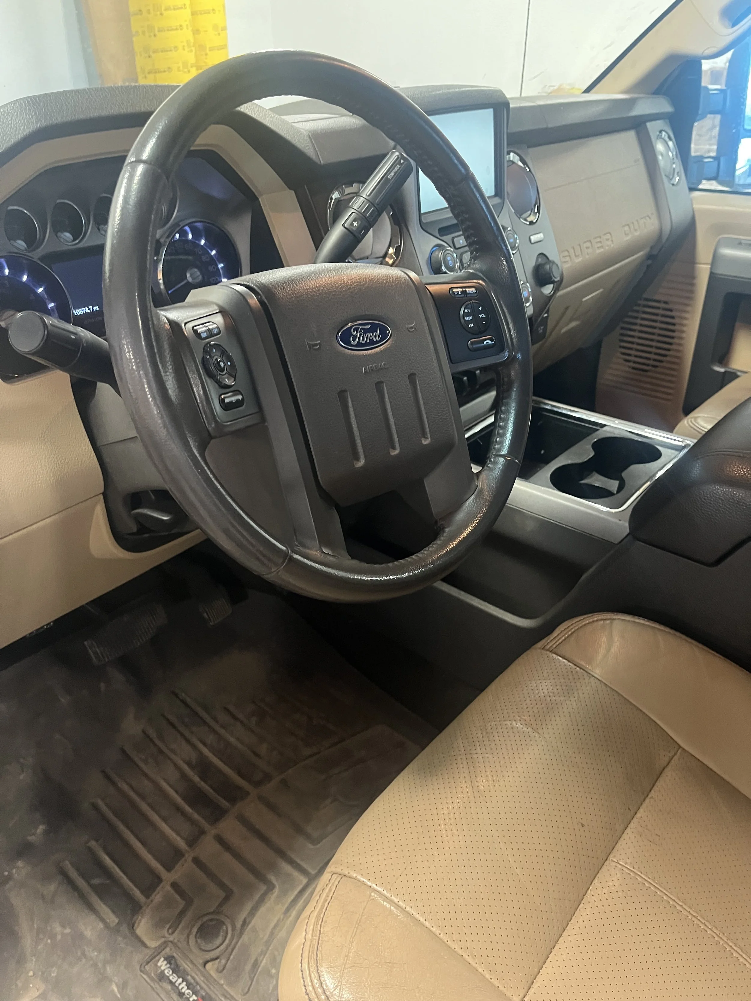 Interior of a Ford vehicle, showing a black steering wheel, dashboard with gauge cluster, center touchscreen display, and beige leather seat with a textured pattern.