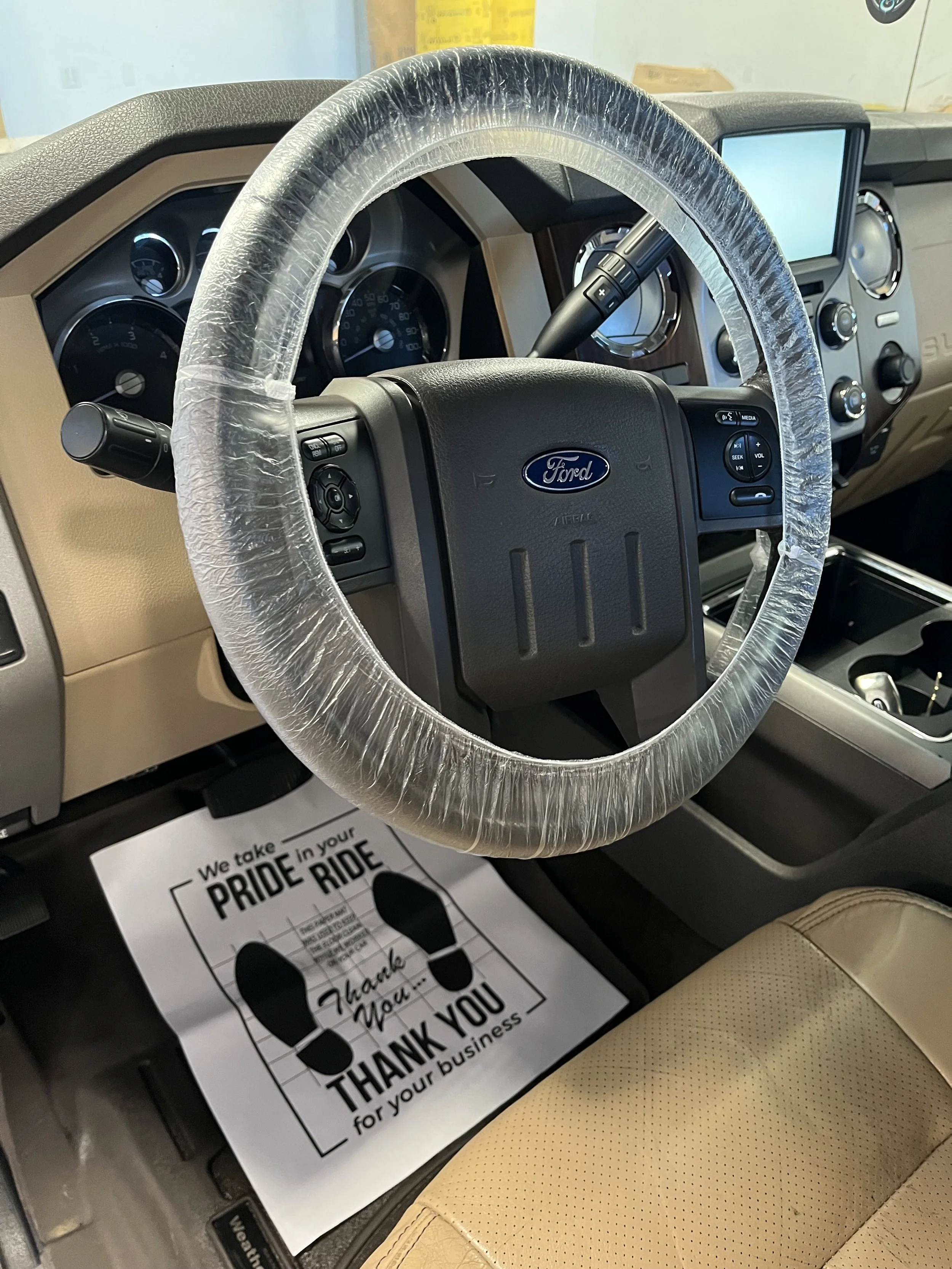 The interior of a Ford vehicle, showing the steering wheel with a plastic cover, dashboard with gauges and controls, and a paper floor mat with a thank you message.