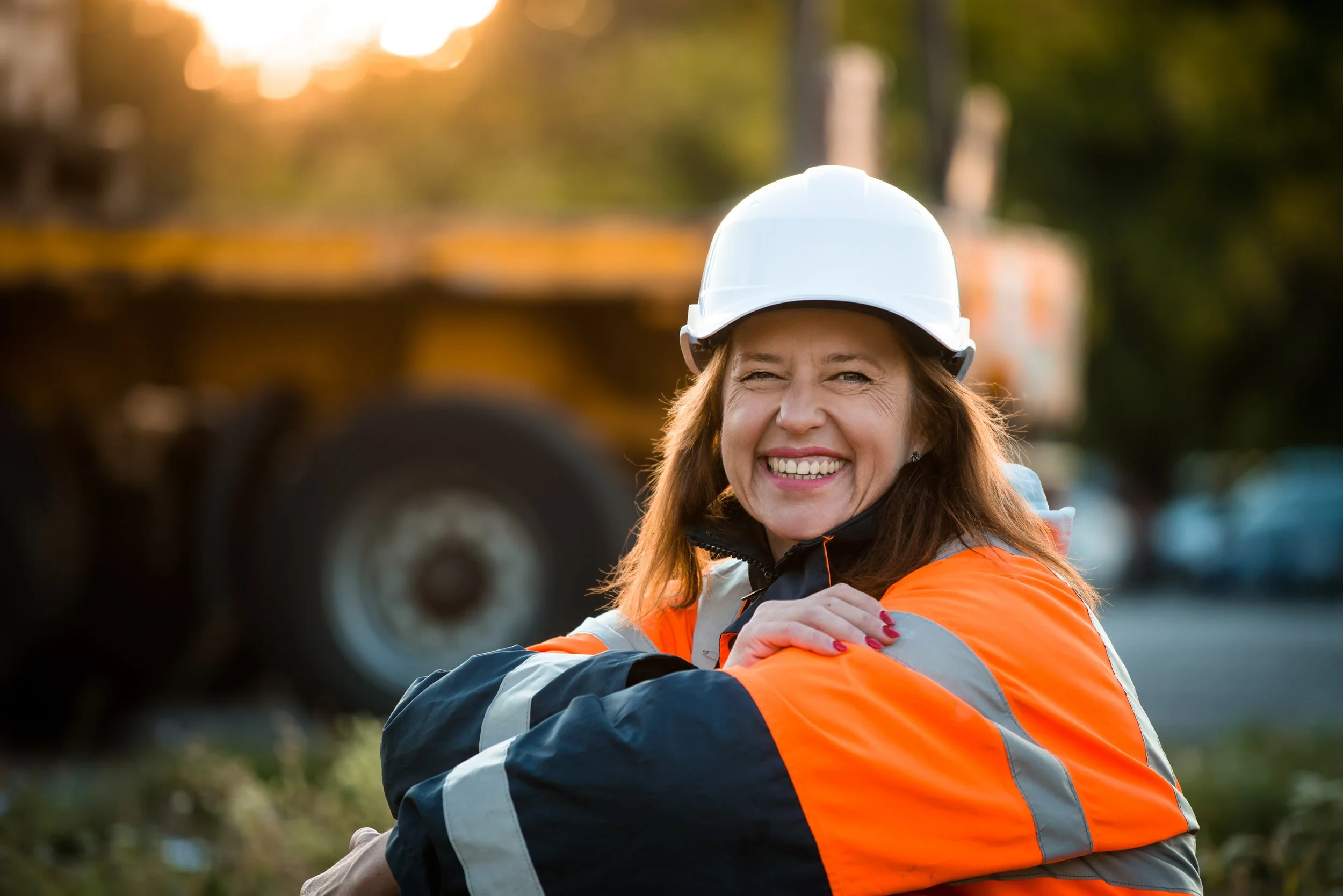 A woman wearing an orange safety jacket and a white hard hat, smiling outdoors with a construction vehicle in the background.
