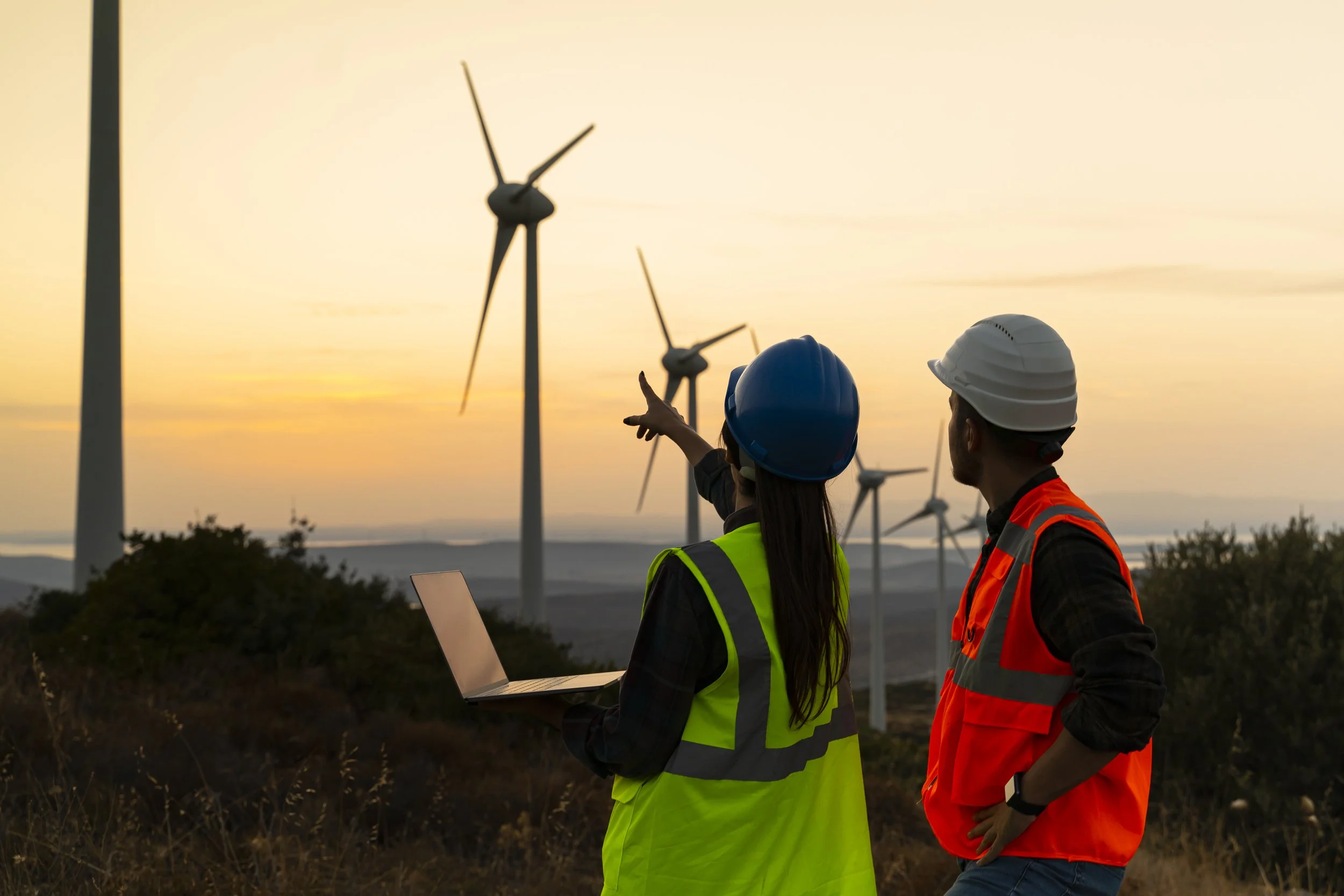 Two wind energy workers in safety vests and helmets consulting at wind turbines during sunset.