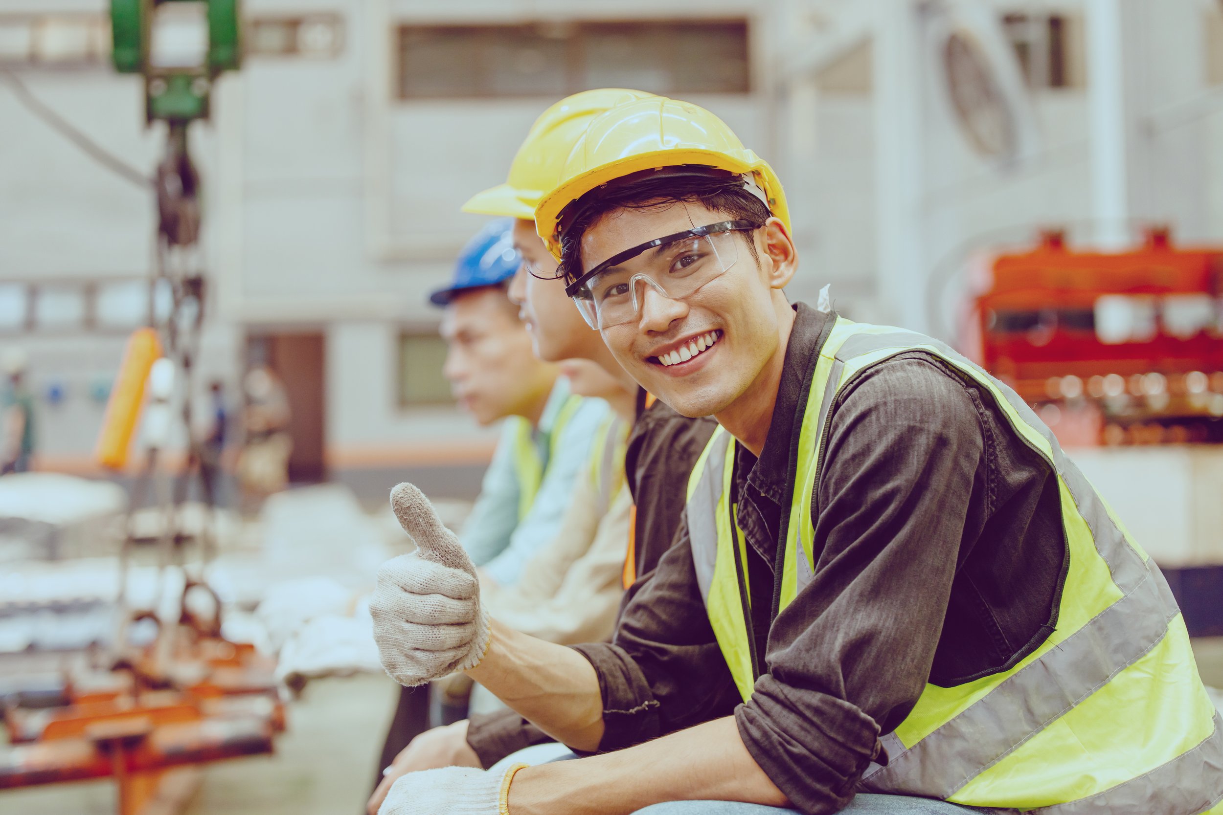 Smiling construction worker wearing a yellow safety helmet and safety glasses giving a thumbs up on a construction site.